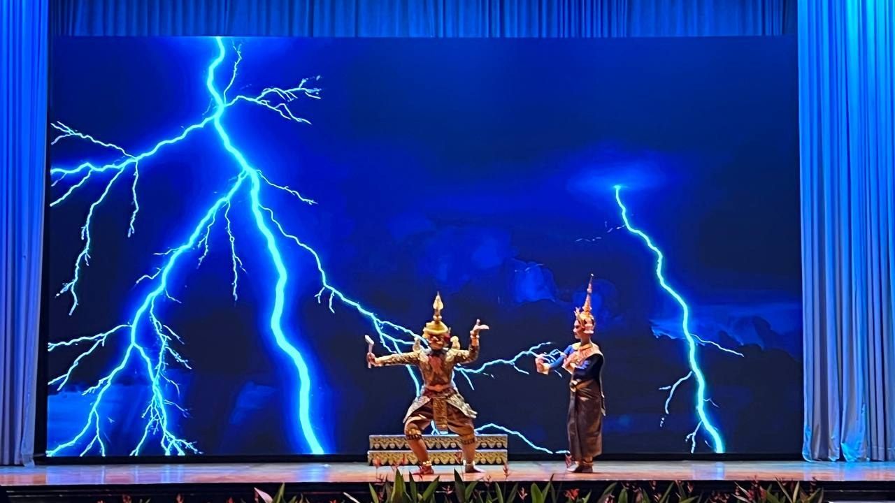 A masked giant and a crowned goddess dance before a stage screen filled with lightning