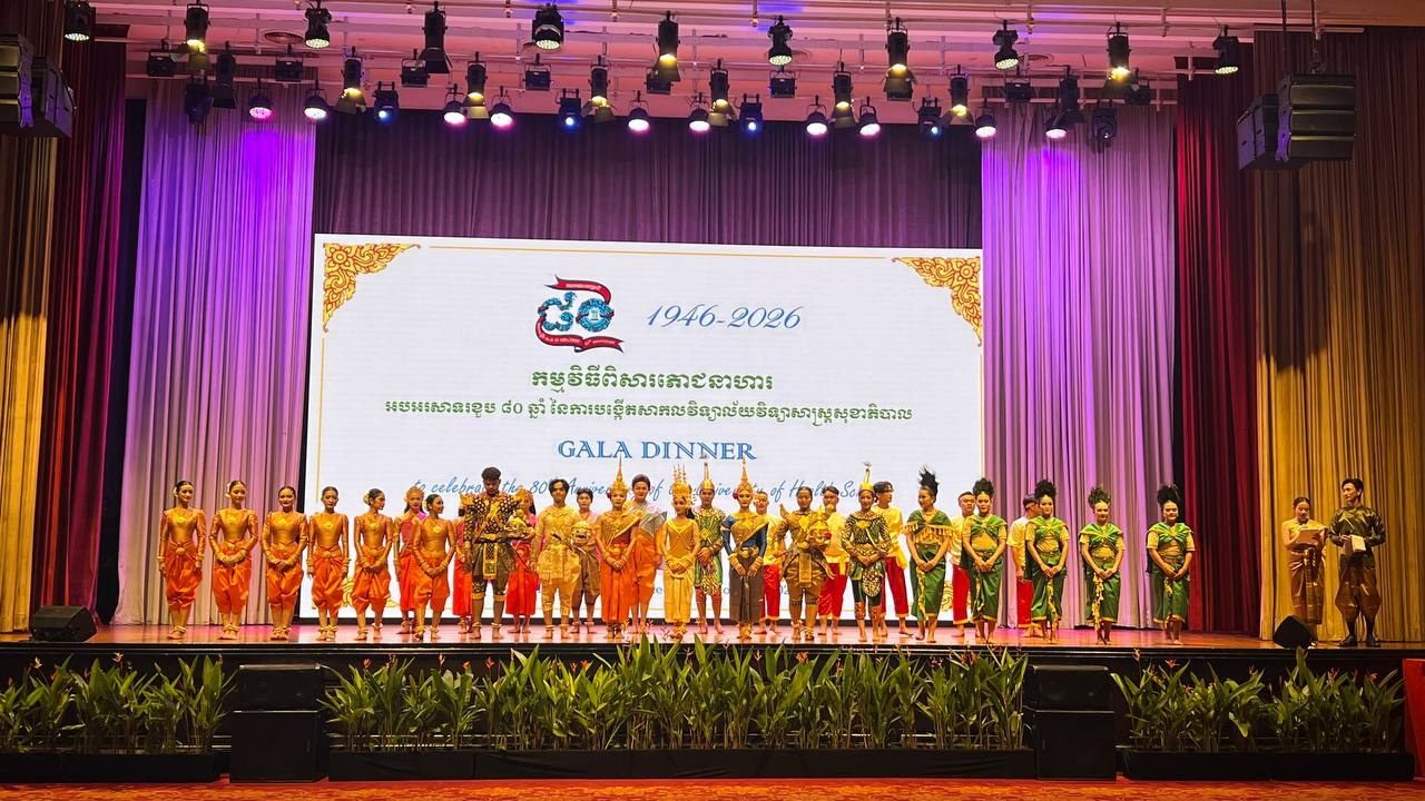 The performers of all seven pieces in full costume in a line on stage beneath the 1946–2026 gala dinner screen, with the two MCs at the right