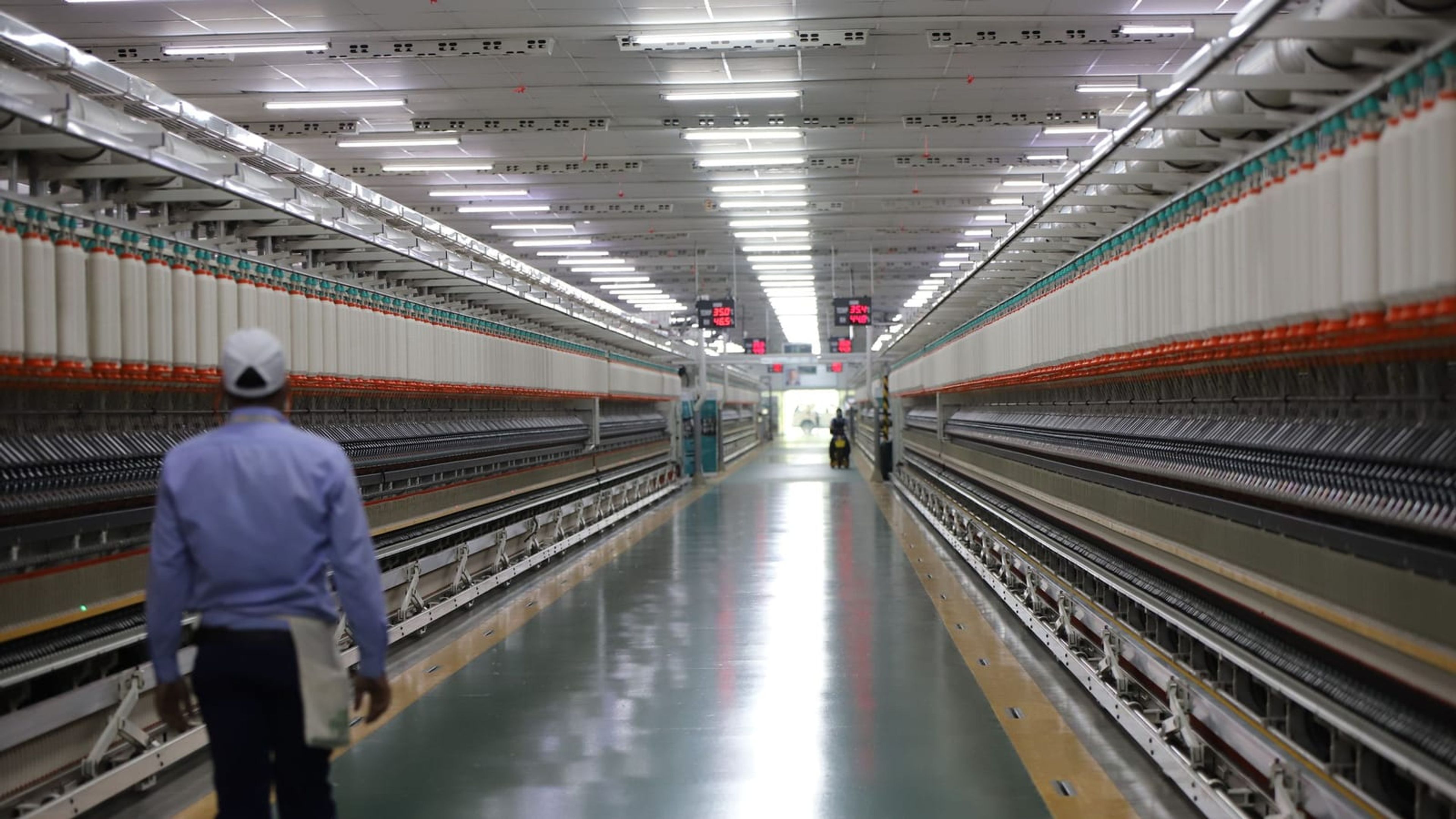 Corredor de fábrica com iluminação LED e técnico caminhando entre as linhas de produção.