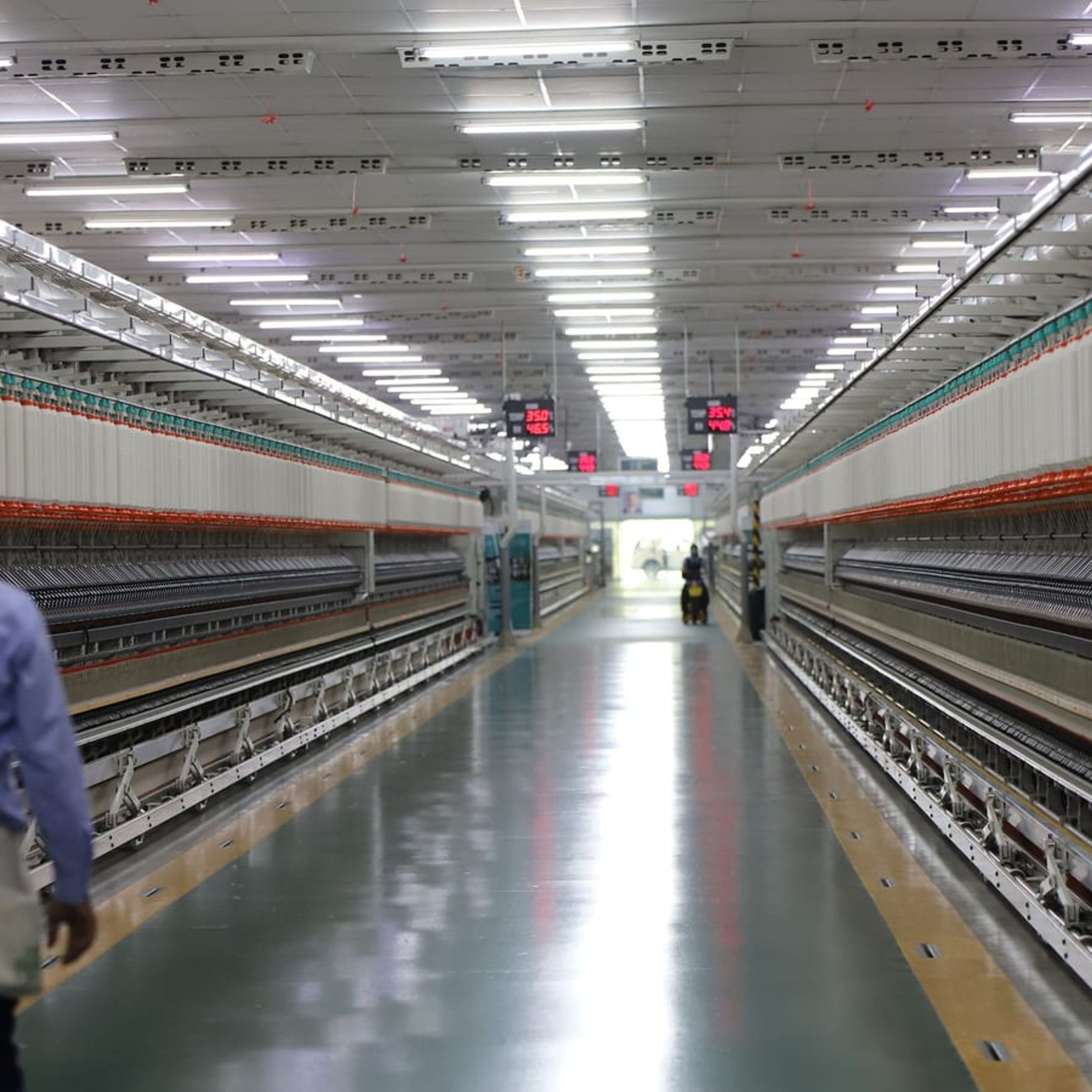 Corredor de fábrica com iluminação LED e técnico caminhando entre as linhas de produção.
