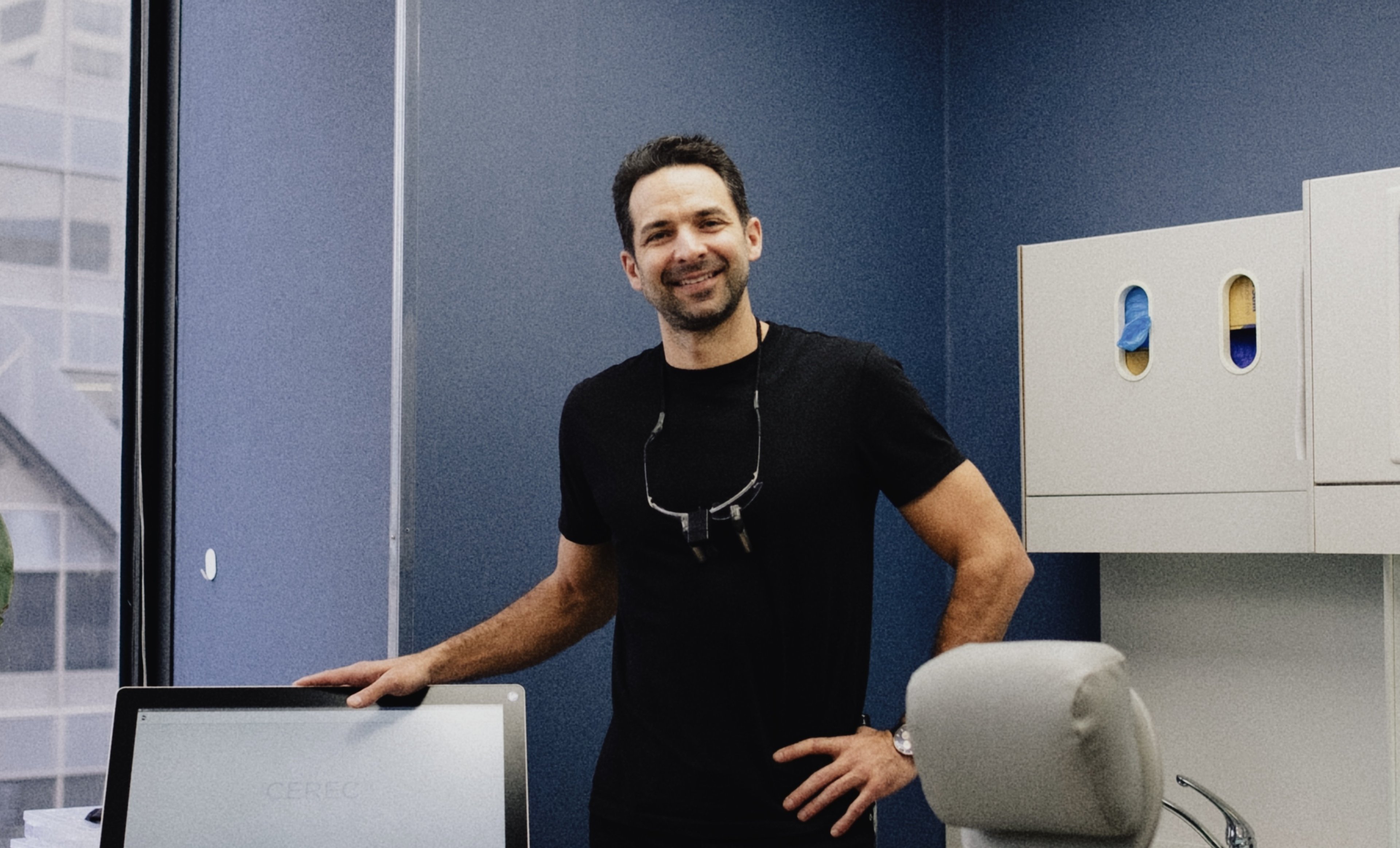 Dr. Louis Santini standing beside a treatment chair, loupes around his neck, smiling.