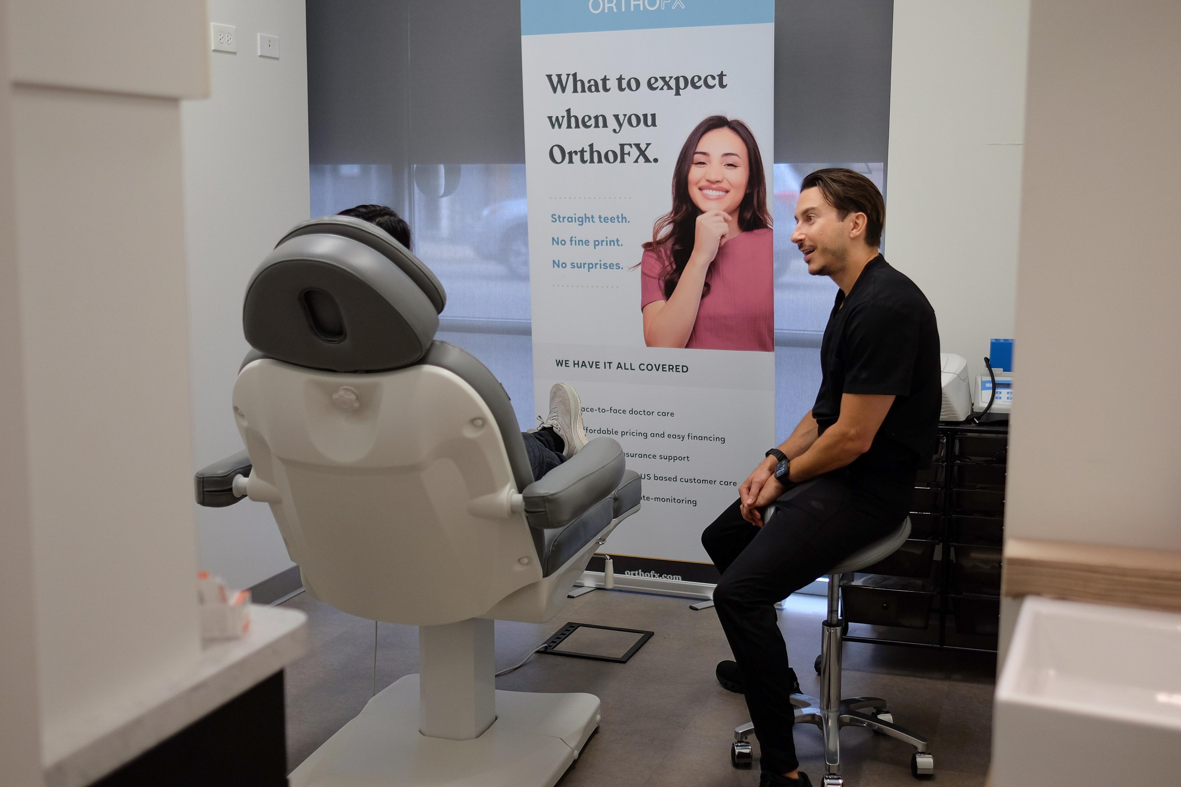 Dr. Vlad Crutcher seated on a stool talking with a patient reclined in a dental chair, an OrthoFX banner standing behind them.