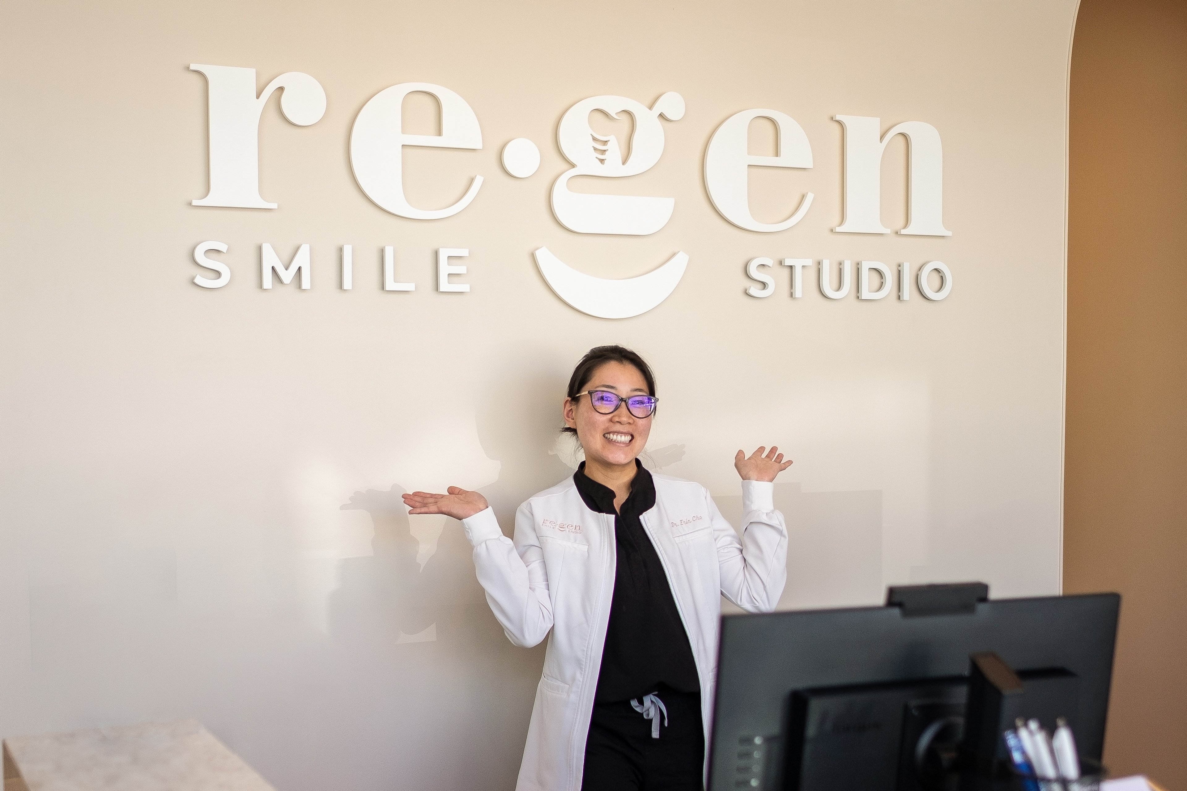 Dr. Erin Cho in a white coat, arms raised, smiling in front of the re·gen smile studio sign.