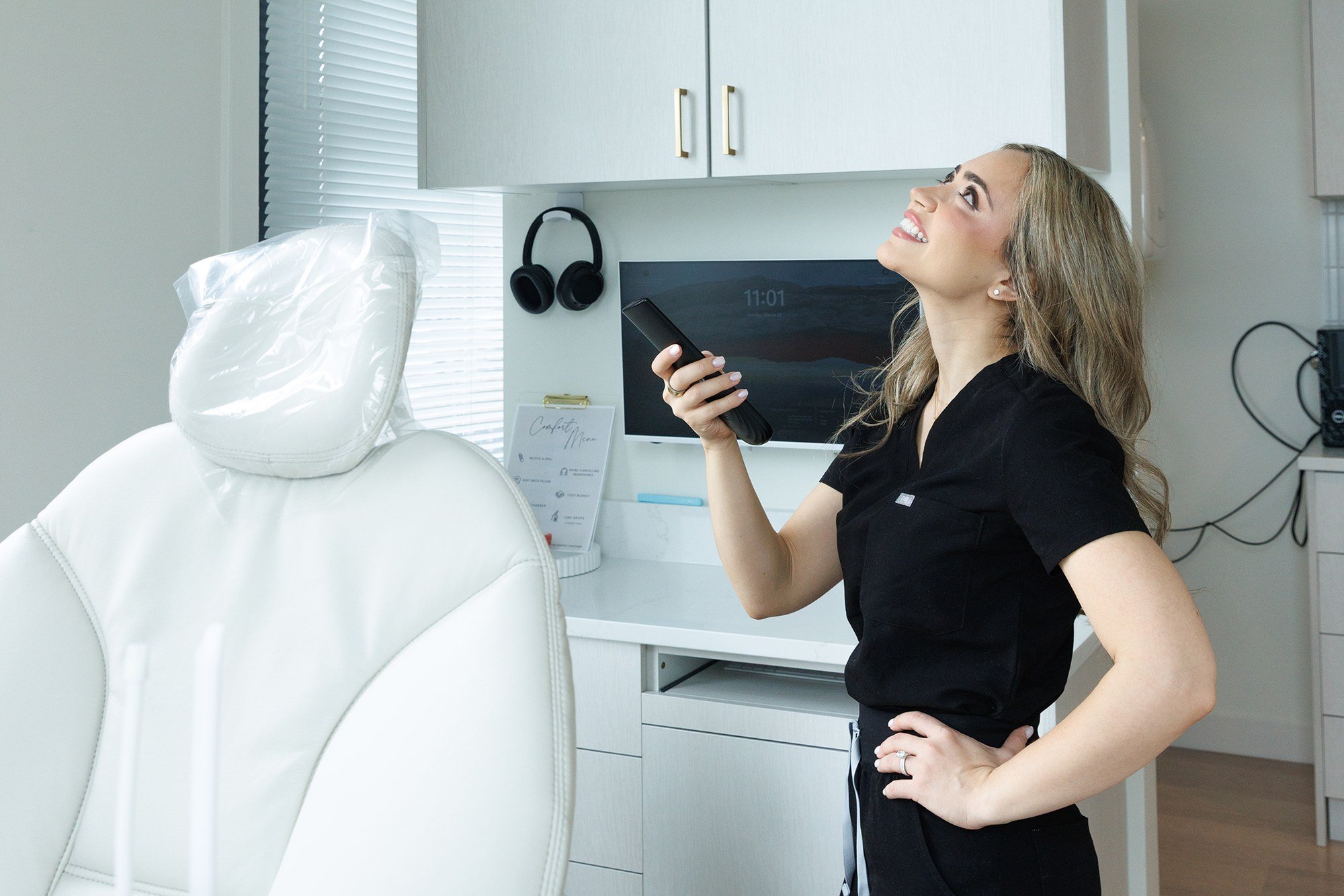 Dr. Renee Alhakim in black scrubs pointing a remote at the ceiling screen in a treatment room, a white dental chair and noise-canceling headphones beside her.