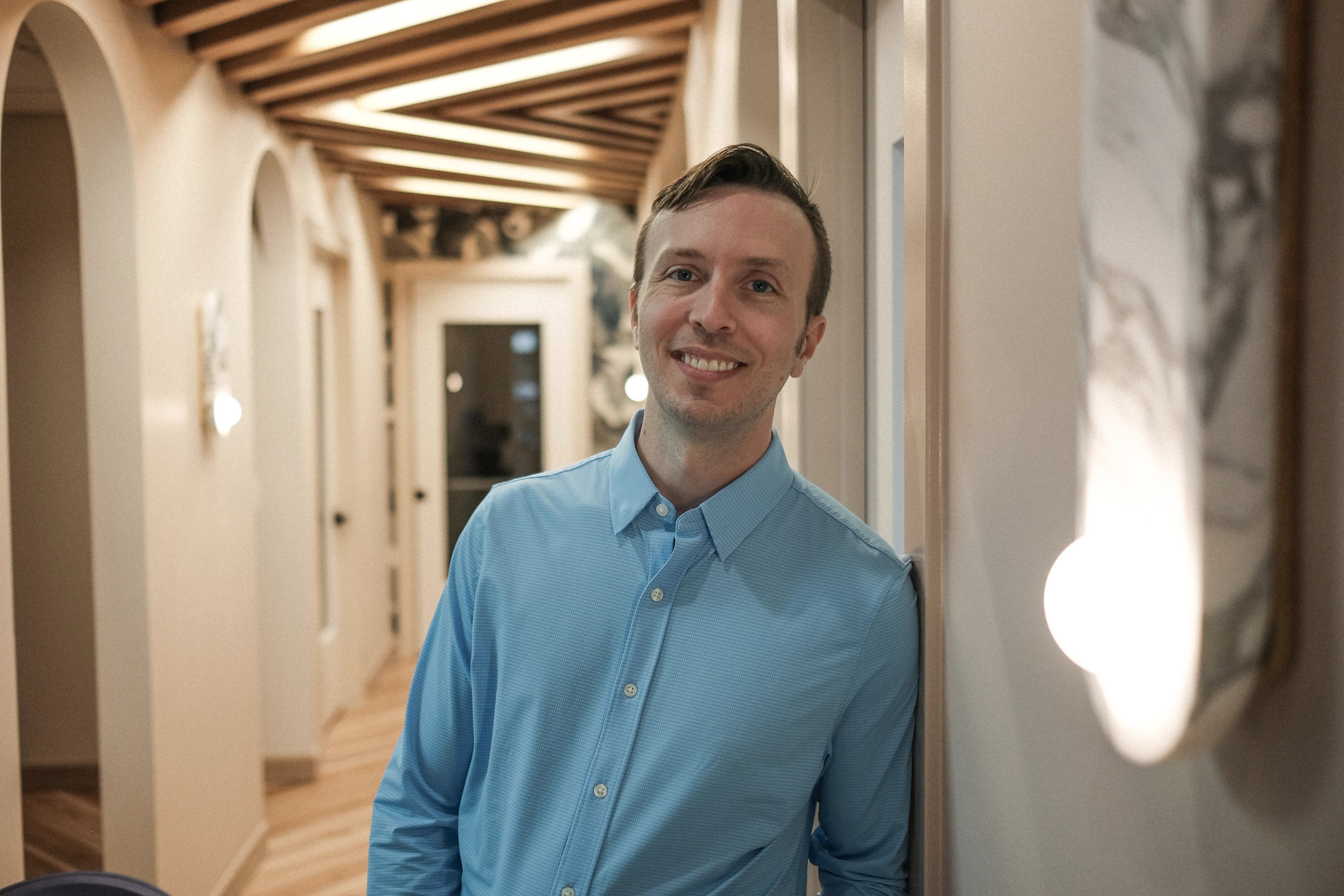 Dr. Ryan Woitas standing in the hallway at Lumiere Dental, leaning against a wall beneath a beamed wood ceiling with warm lights running down the corridor.
