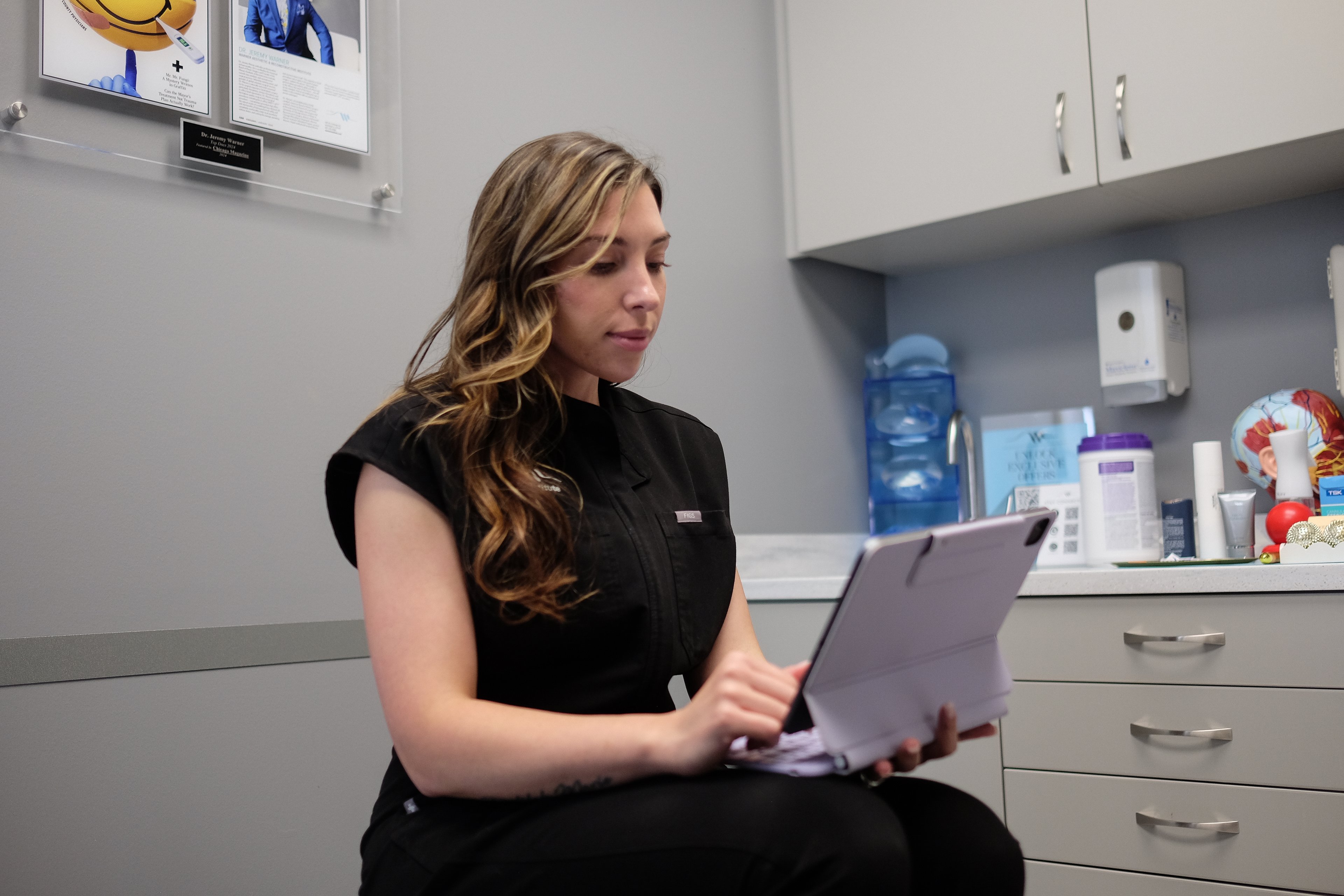 Alyssa Nasman in black scrubs looking down at a tablet in a treatment room at the Warner Institute, cabinets and supplies on the counter behind her.