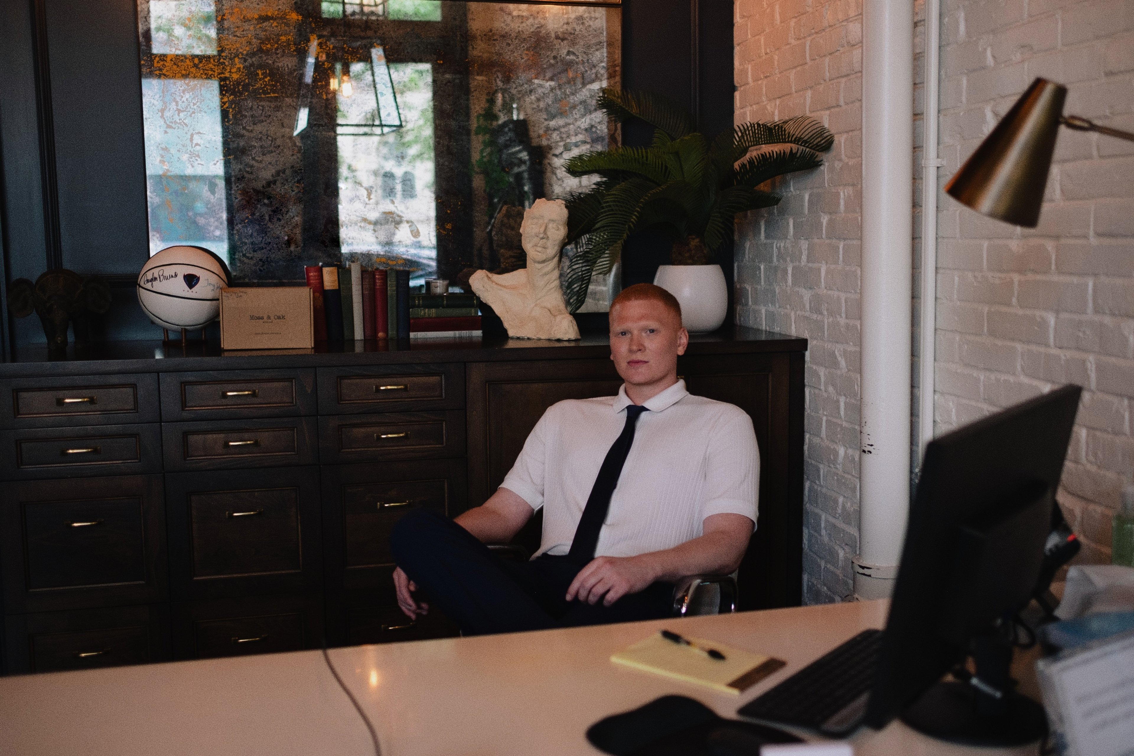 Dr. Colby James Gerth seated at a desk at Pearl in a white shirt and dark tie, a dark wood cabinet, plaster bust, and mirror behind him.