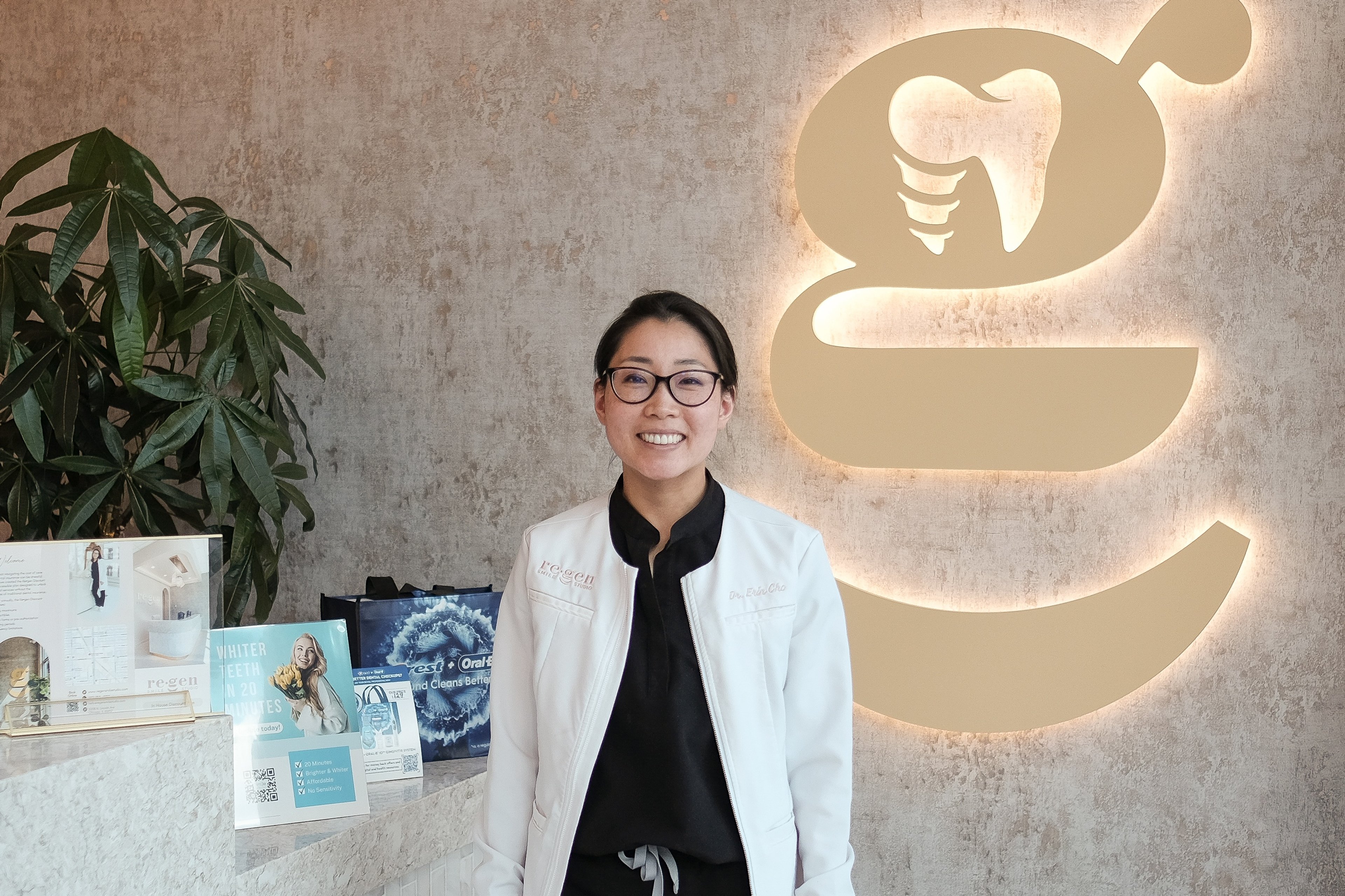 Dr. Erin Cho standing in the lobby of her practice in front of a large illuminated lowercase g letter on a concrete wall, a tooth set into the letter.