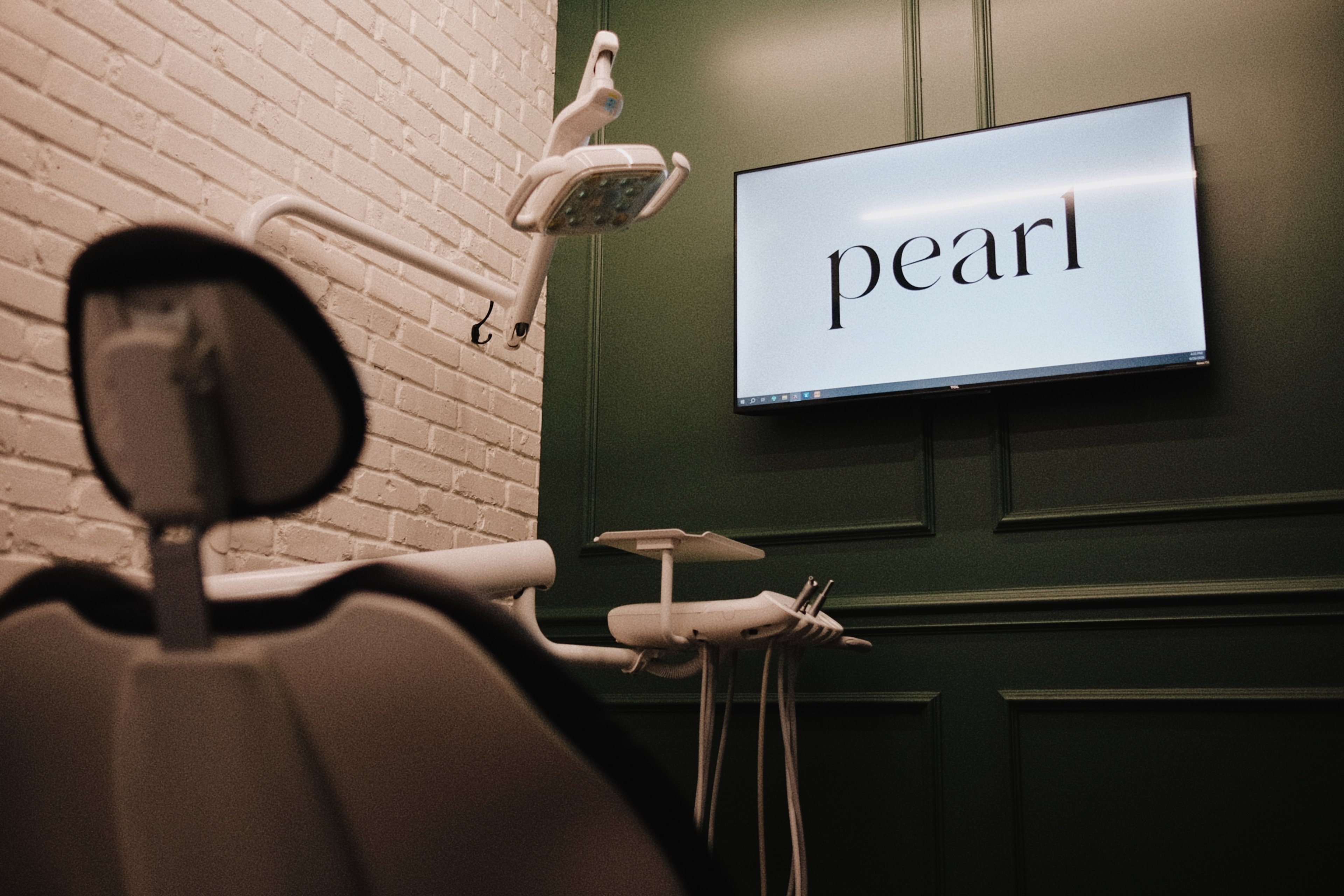 A treatment room at Pearl with a dark dental chair, white brick, and a screen reading pearl on a green wall.