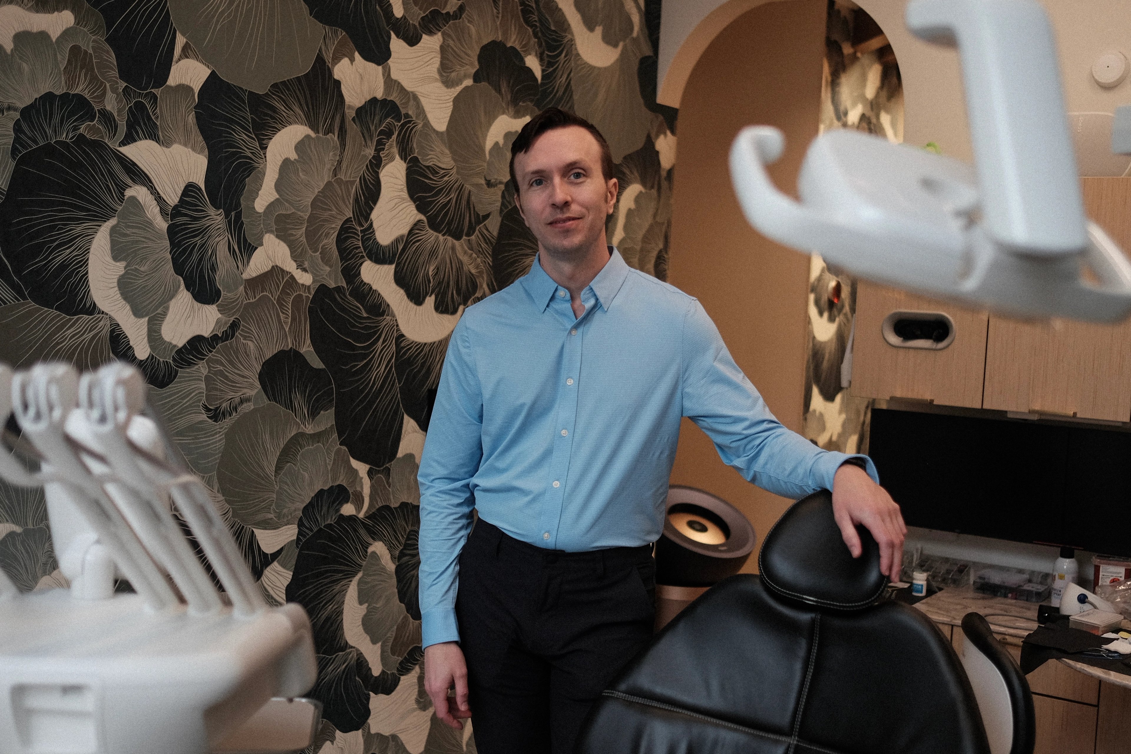 Dr. Ryan Woitas standing beside a dental chair in an operatory at Lumiere Dental, in front of a large floral patterned wall.