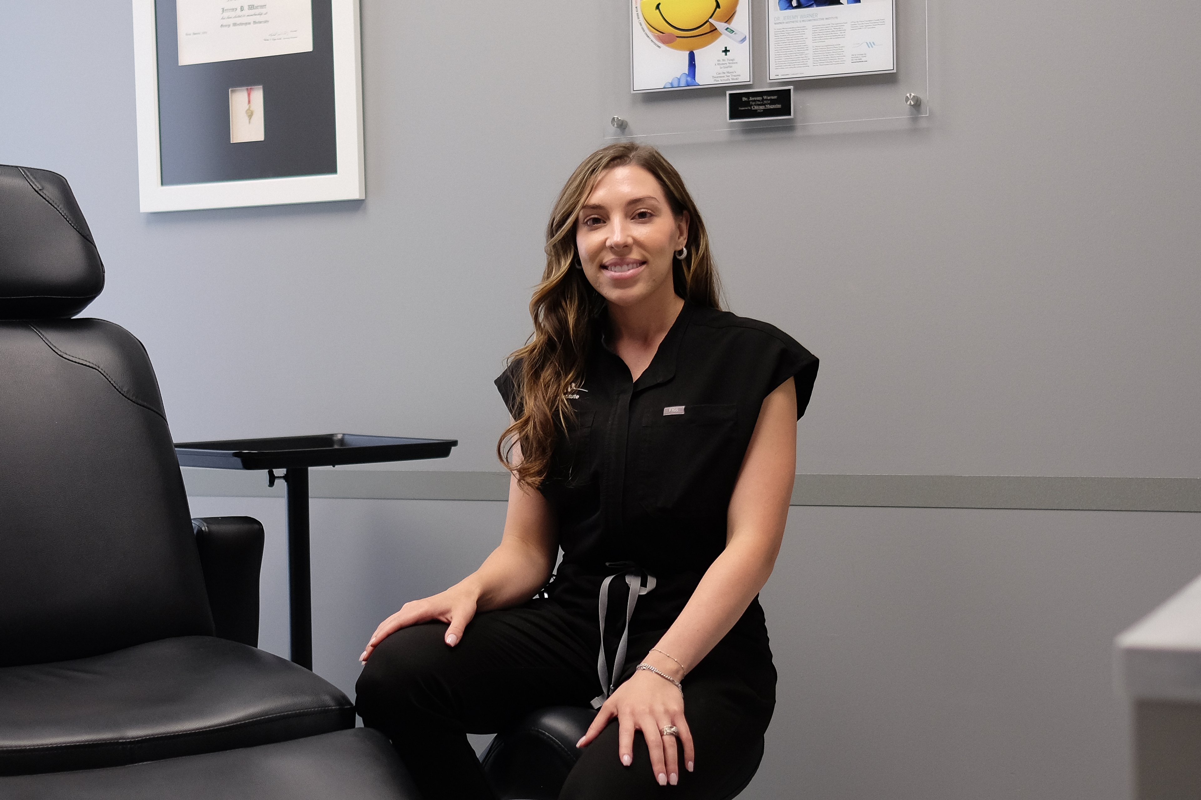 Alyssa Nasman seated beside the black treatment chair in her gray treatment room at the Warner Institute, framed certificates on the wall behind her.