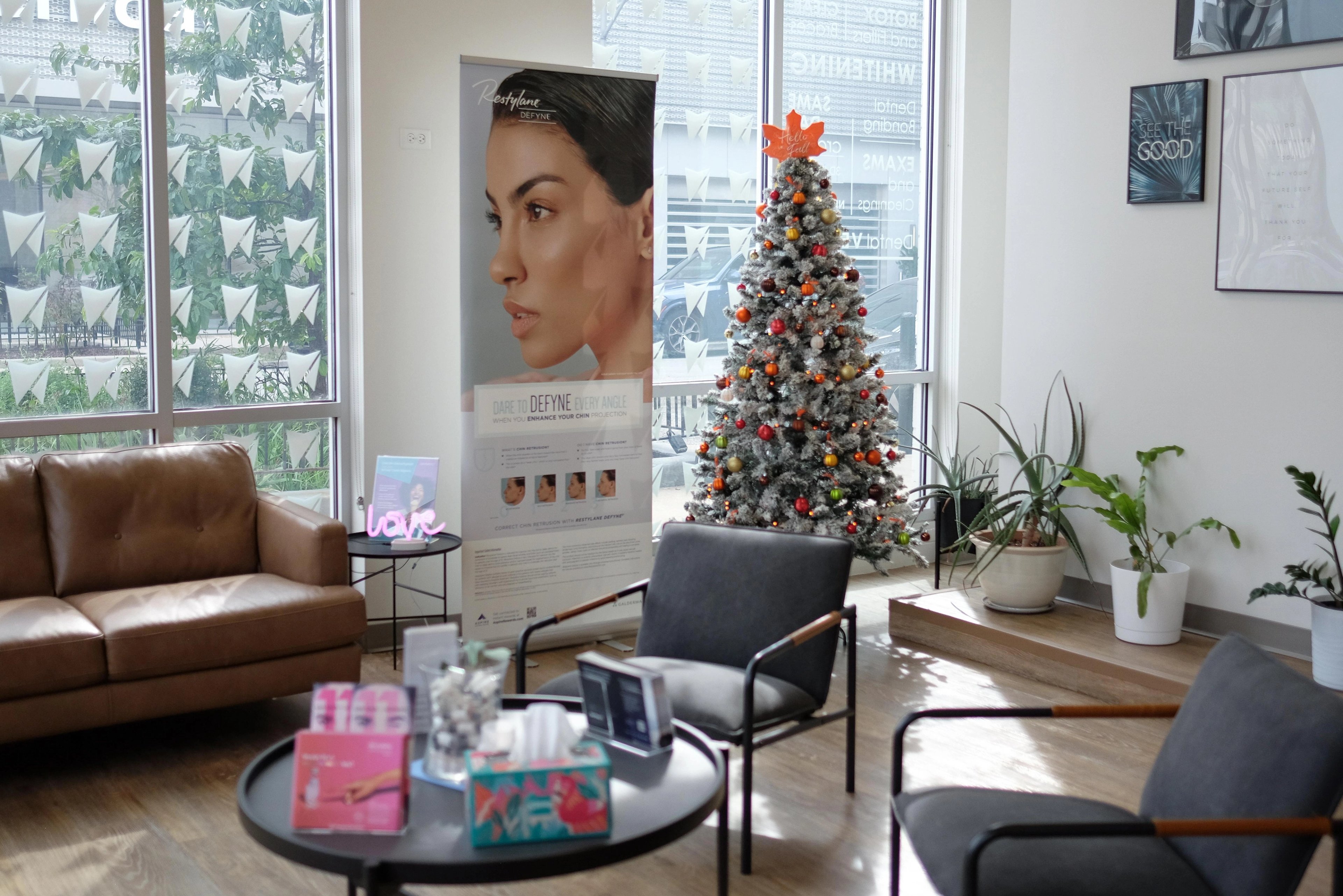 The waiting room at The Dental Standard, a white-dusted tree hung with orange, red, and gold ornaments and topped with a maple leaf standing by the front windows beside a leather sofa and gray armchairs.
