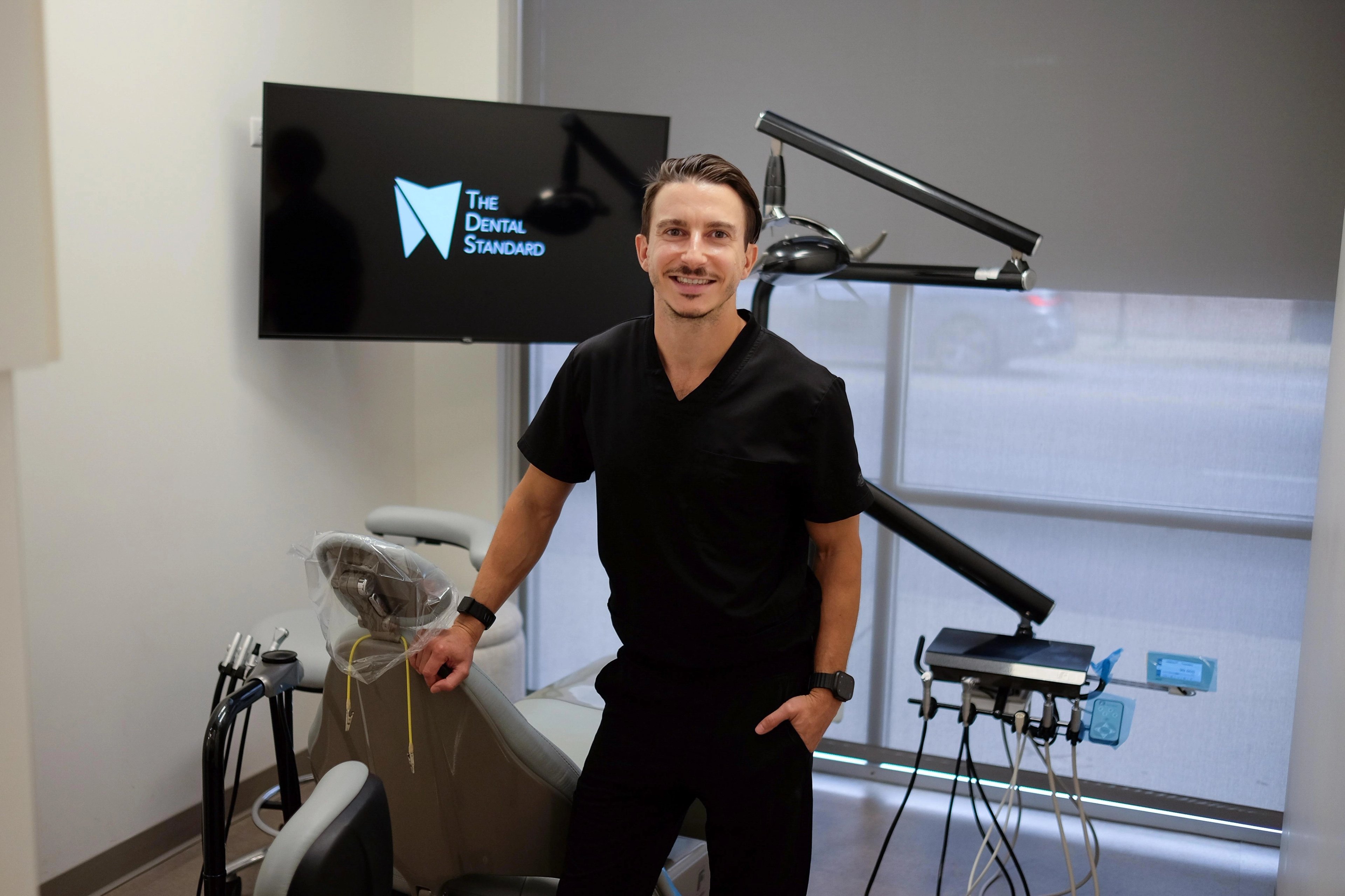 Dr. Vlad Crutcher in black scrubs leaning on a dental chair in his treatment room, a wall screen showing The Dental Standard logo behind him.