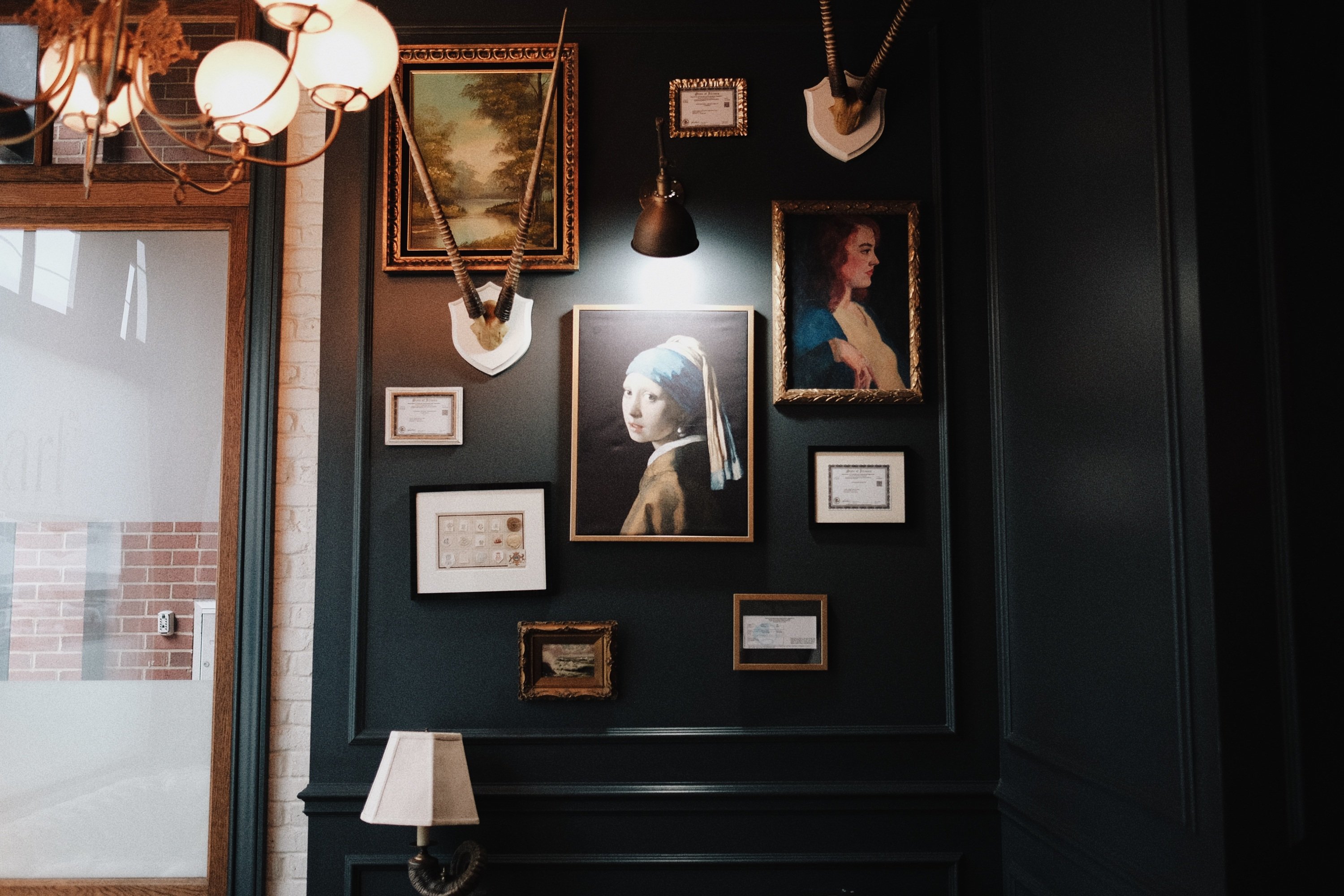A dark green wall at Pearl hung with mounted horns, small gilt-framed pictures, a lamp, and the Girl with a Pearl Earring at the center.