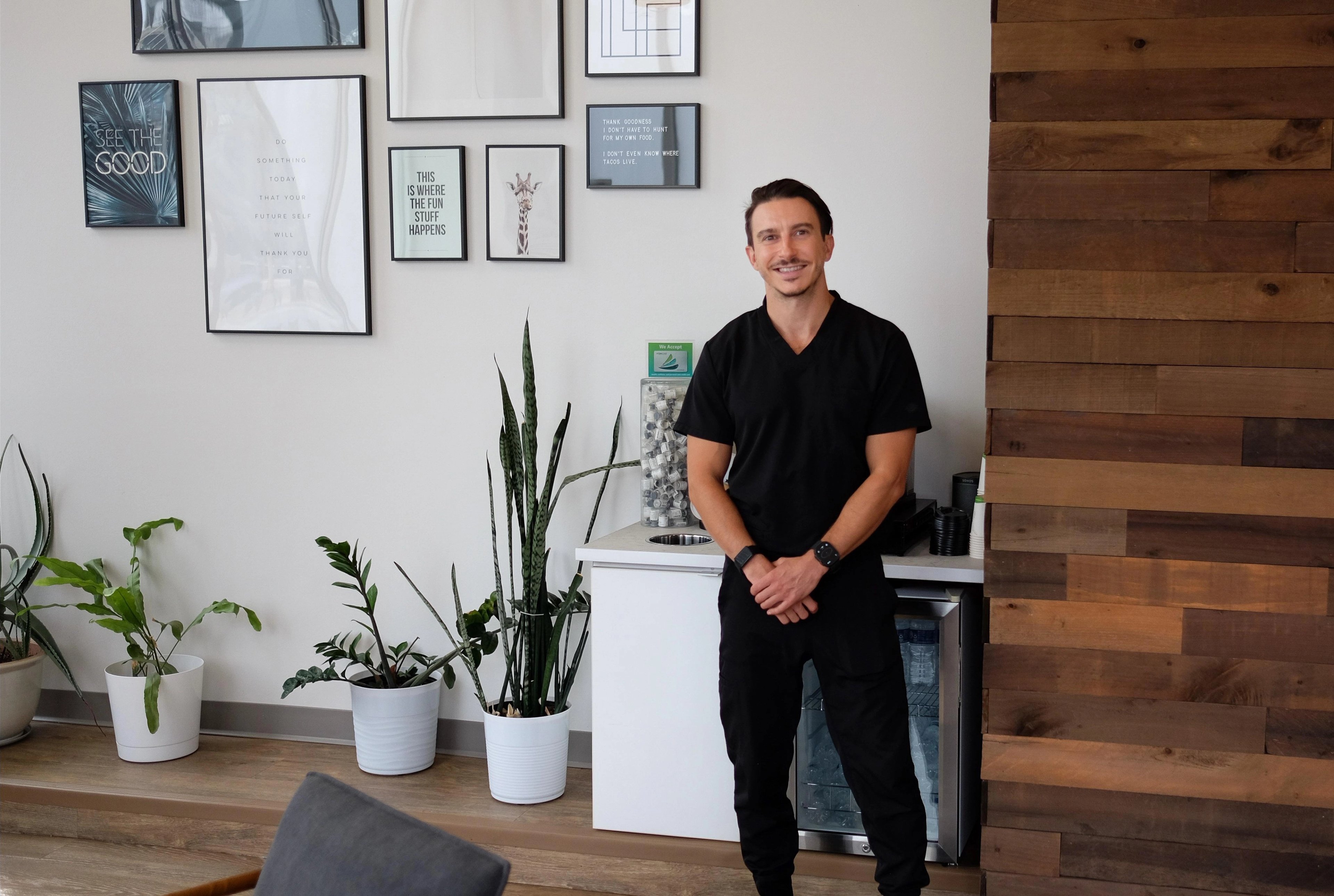 Dr. Vlad Crutcher in black scrubs standing with his hands clasped in the waiting room of The Dental Standard, a gallery wall of framed prints and potted plants beside him and a reclaimed wood wall to his left.