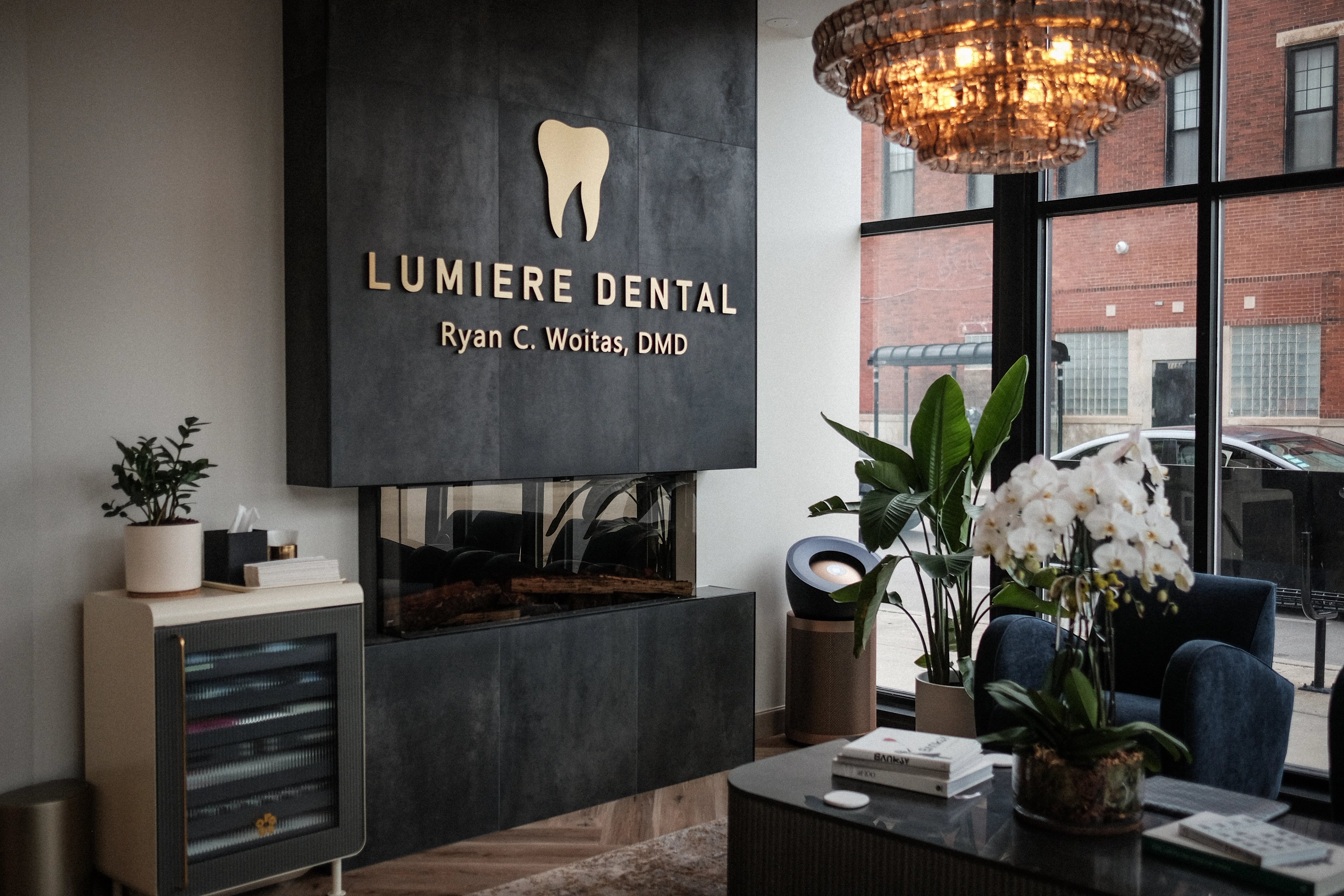 The reception at Lumiere Dental, a dark stone desk under a gold tooth logo and the practice name, with a lit chandelier beside the front window.