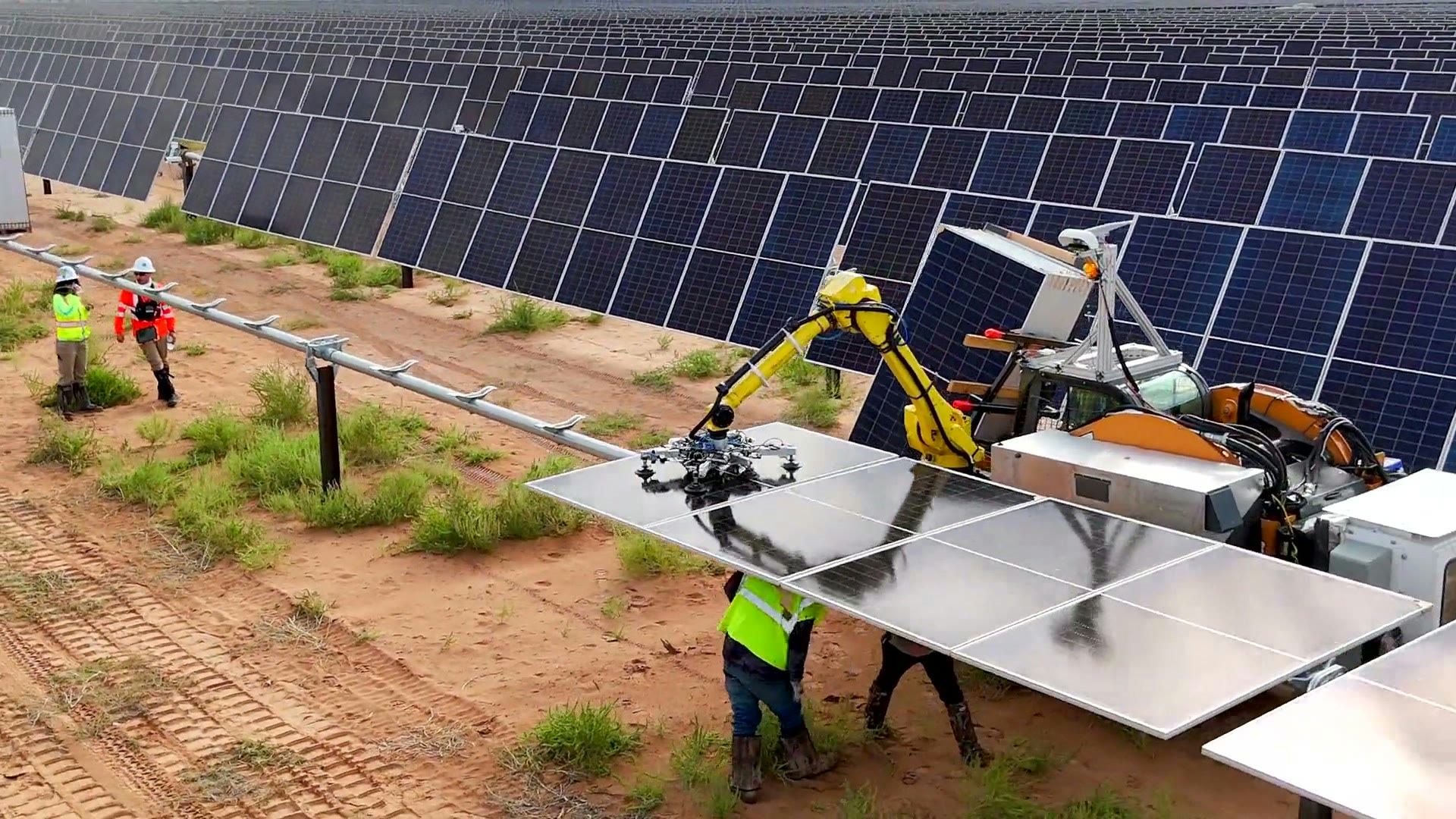 A Gritt robotic arm lowering a solar panel into a row on an active desert solar farm, a worker in safety gear alongside