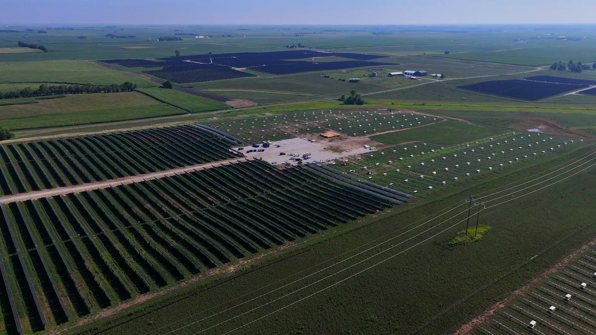 Low aerial over green fields at a solar installation, panel rows on one side and wind turbines on the horizon