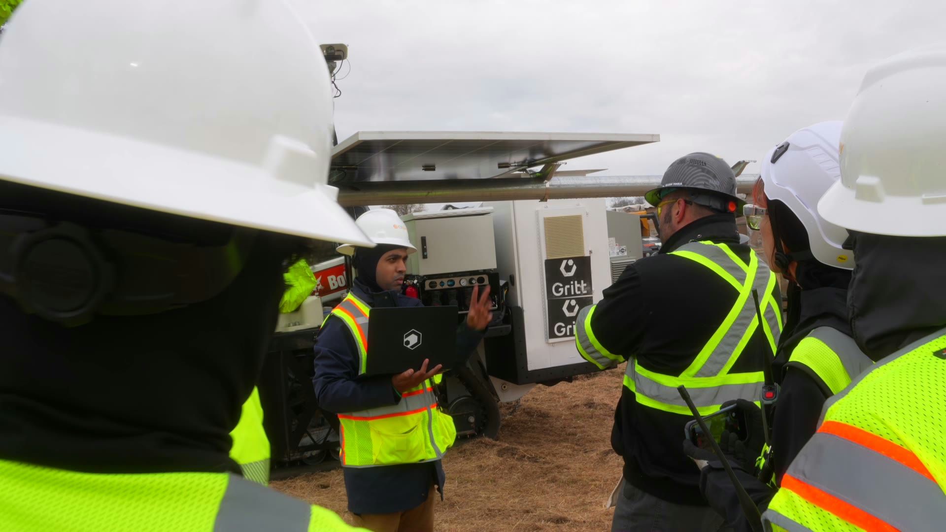 A Gritt crew in hard hats and hi-vis gathered around the machine on a jobsite, one presenting on a laptop