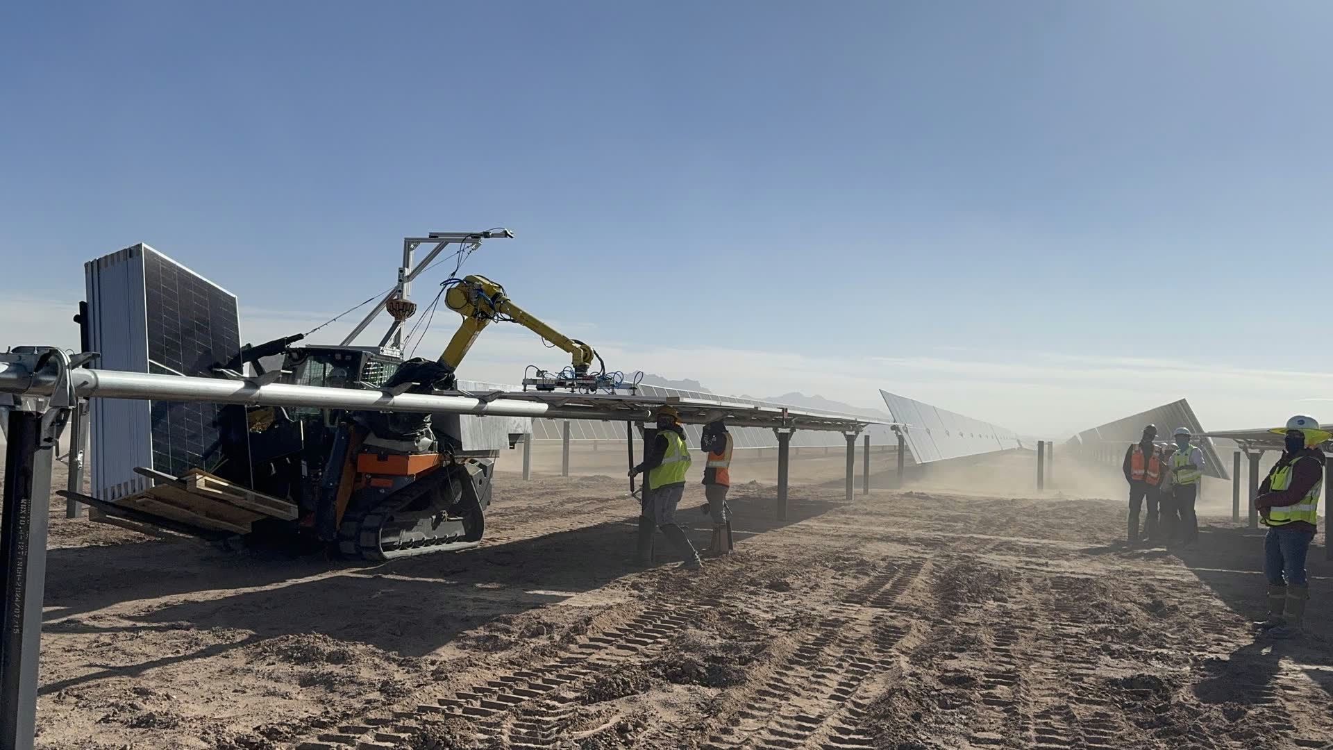 A Gritt machine with a robotic arm working a dusty desert solar construction site, crew in hard hats and safety vests alongside rows of panels