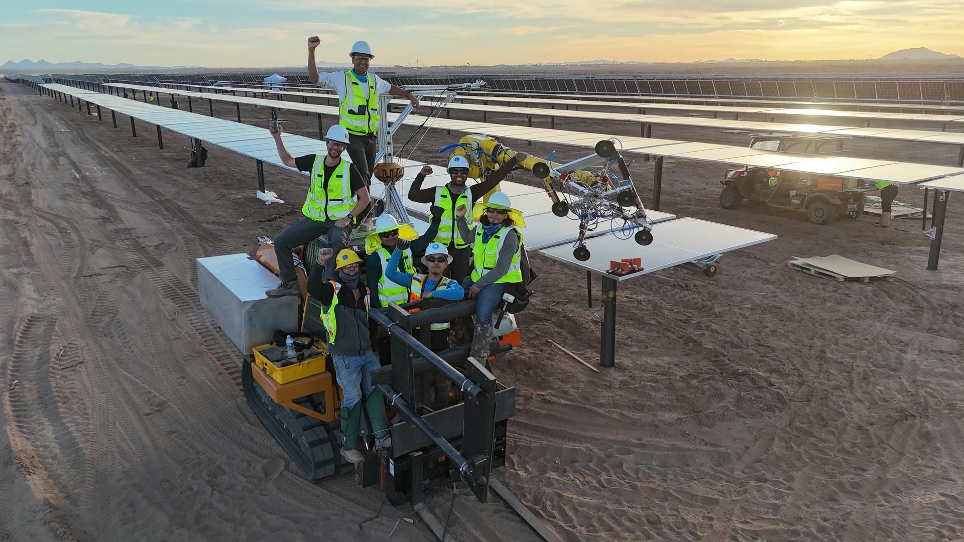 The Gritt crew in hi-vis gathered on their machine at a desert solar installation at sunset, raising fists in celebration, rows of panels stretching to the horizon