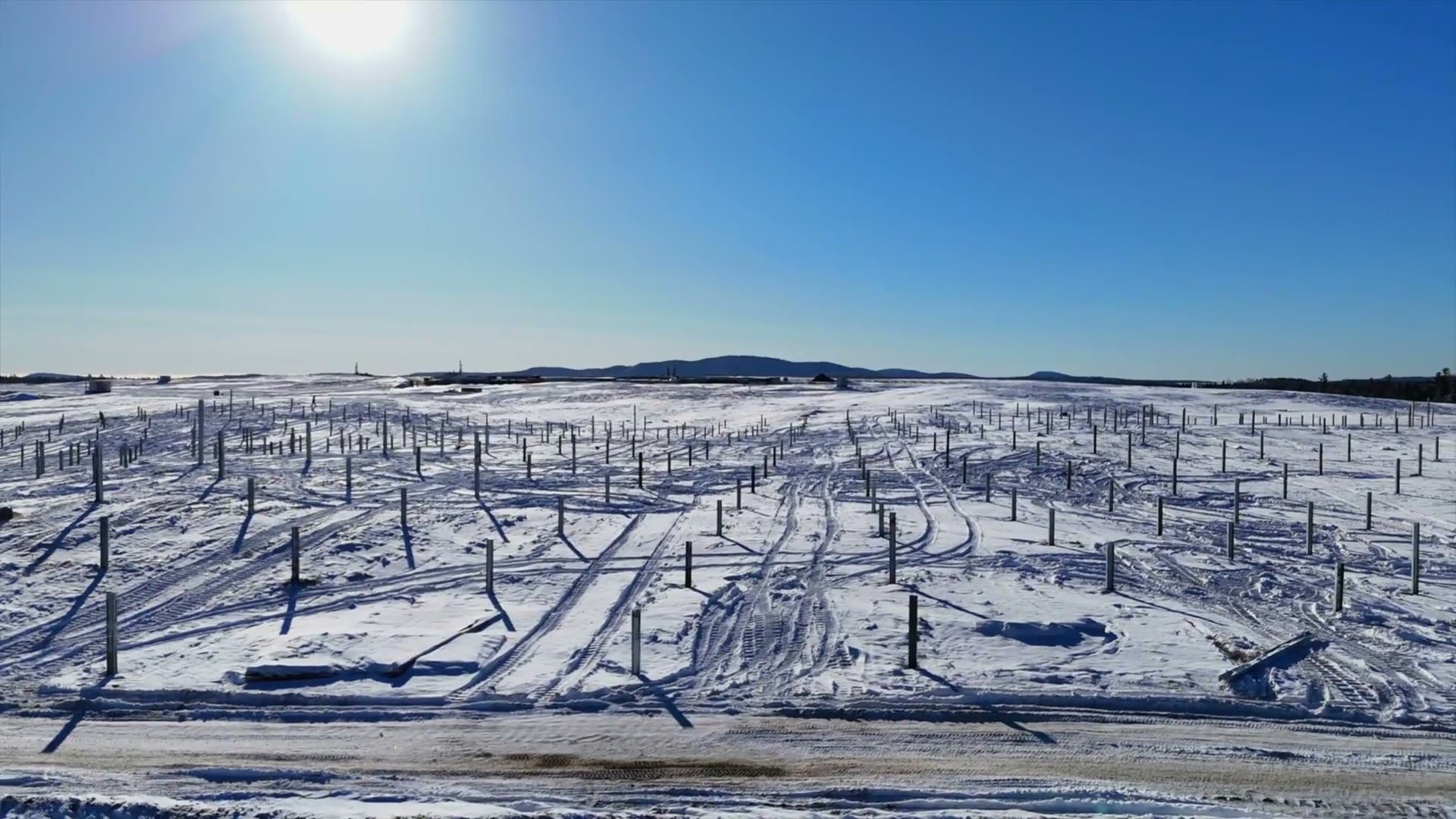 Aerial view of a snow-covered solar construction site, rows of mounting posts across a frozen field under bright sun