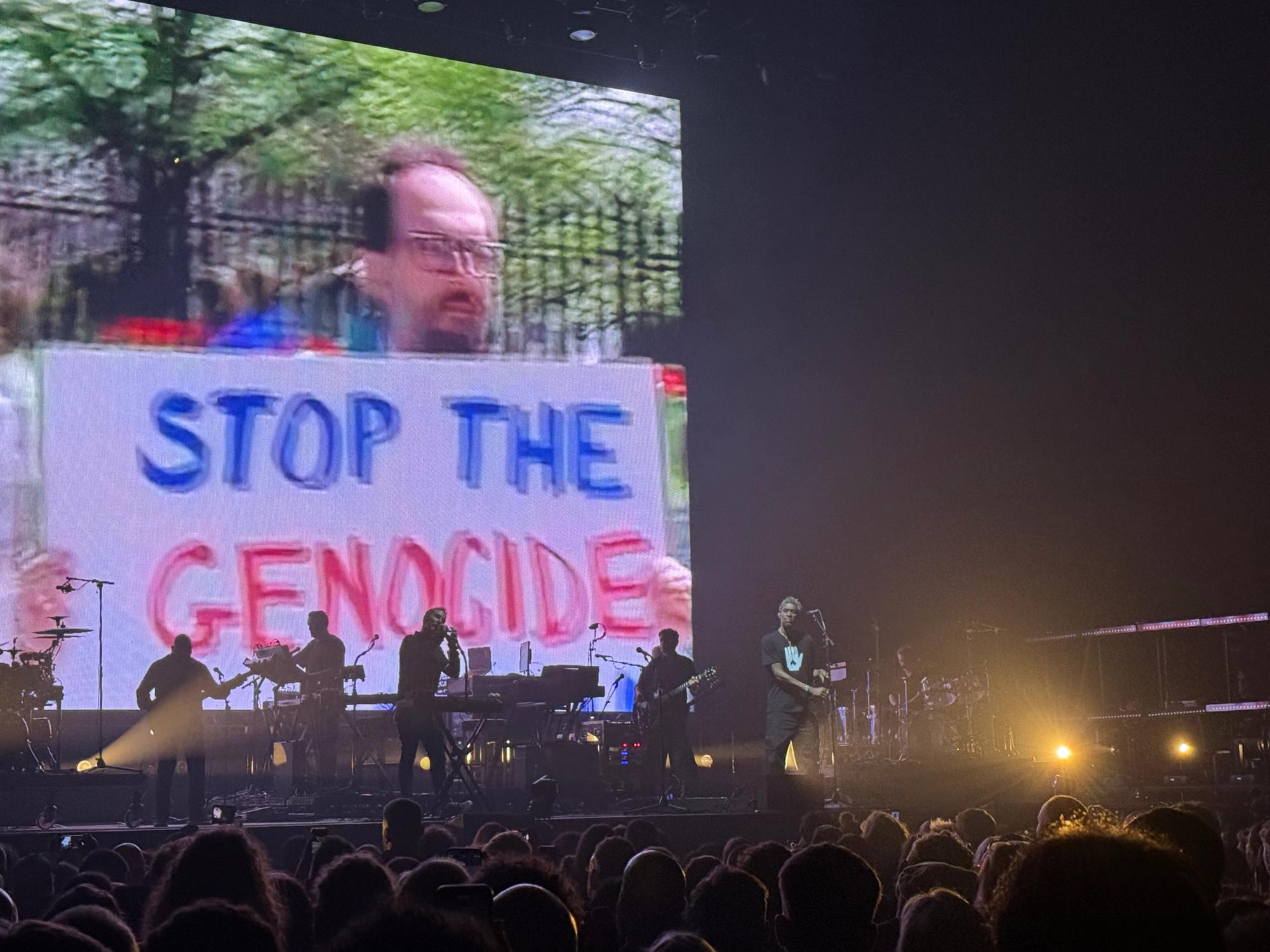Grant at the mic, behind is a picture of someone holding a 'stop the genocide' sign