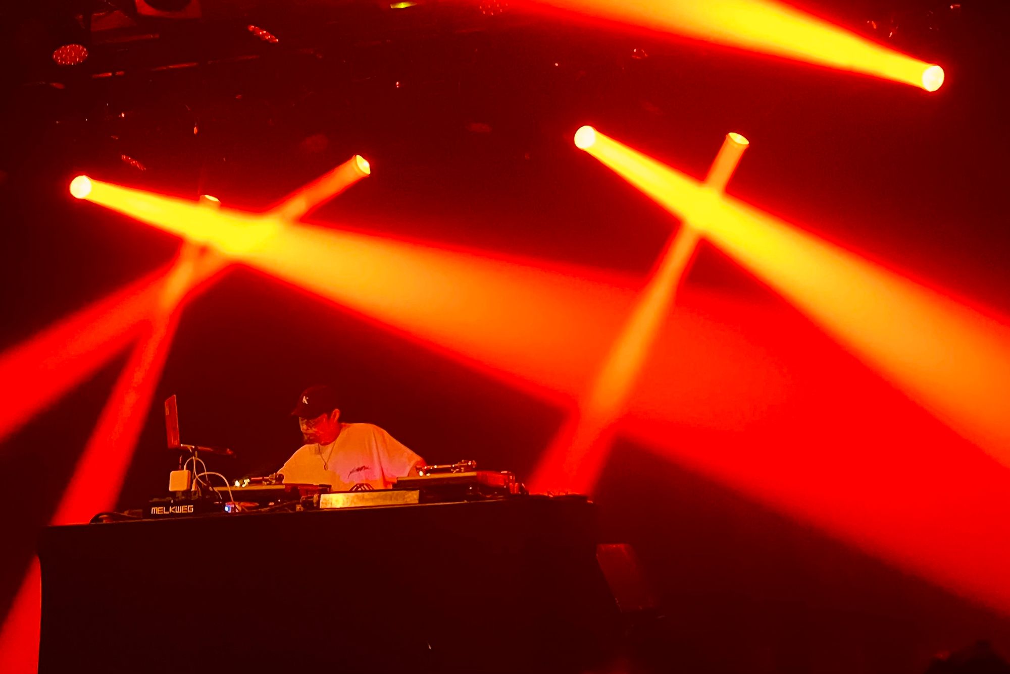DJ Krush behind table with two record players and a laptop, red spotlights against a black background