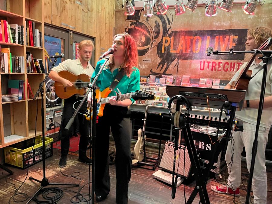 front women Jeanne in the middle singing while playing guitar, with guitarists left and right, in the back is the Plato Live Utrecht logo