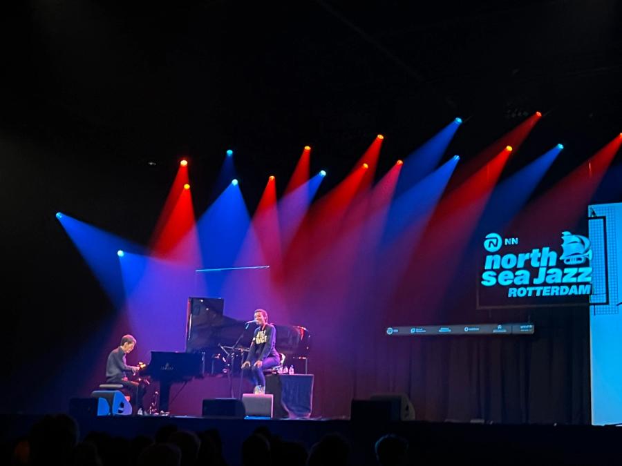 Fred on the left behind piano, Esperanza seated next to him; spotlights are blue and red, on the right is the north sea jazz rotterdam logo with a ship that is also a trumpet
