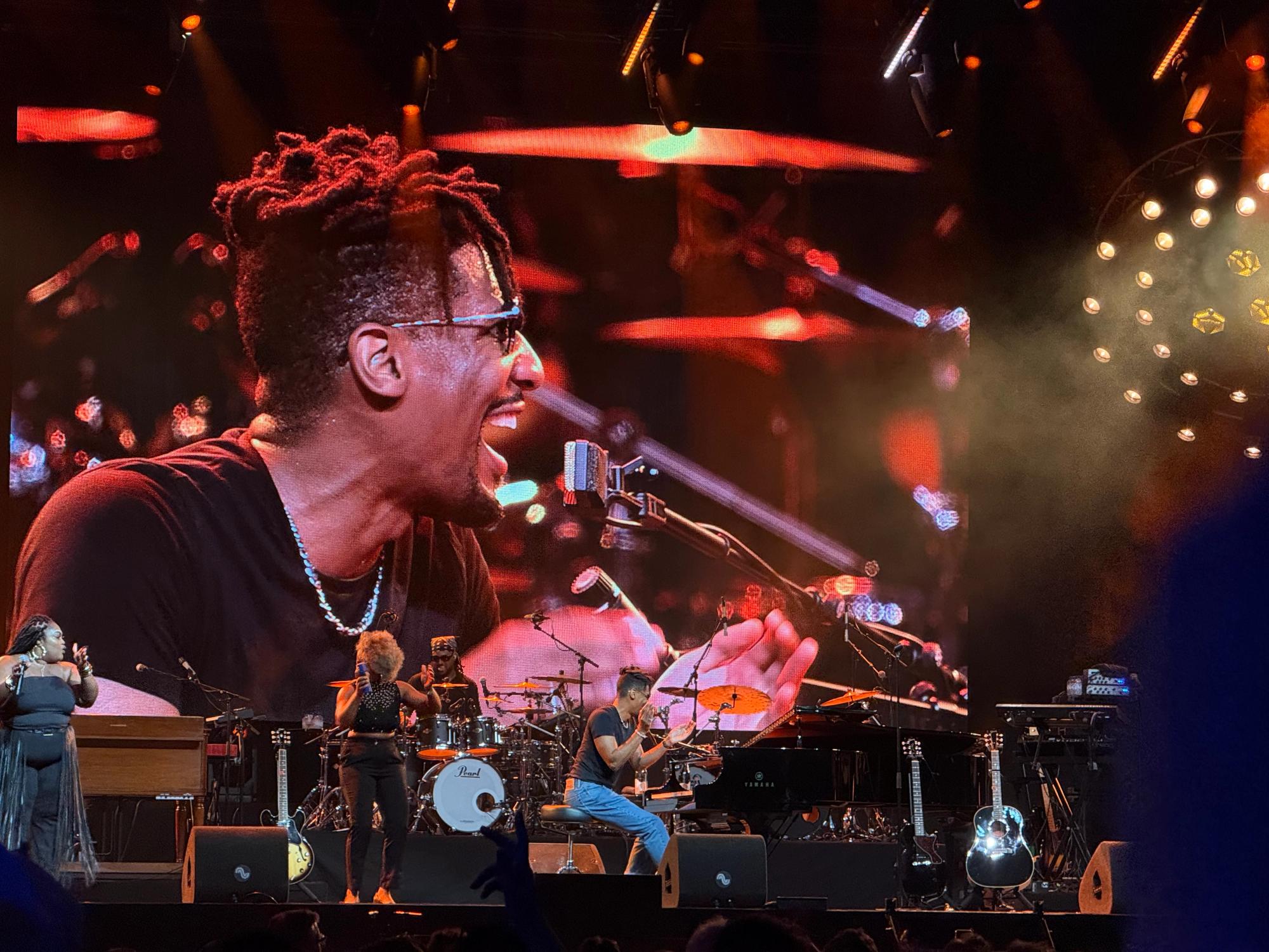 Jon Batiste playing piano with at the back a huge video screen that shows him playing and laughing