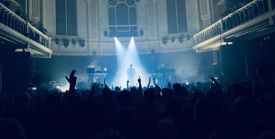 view of stage from crowd; on stage there is the shadow of person covered in lights, on the right a piano player and on the left a DJ