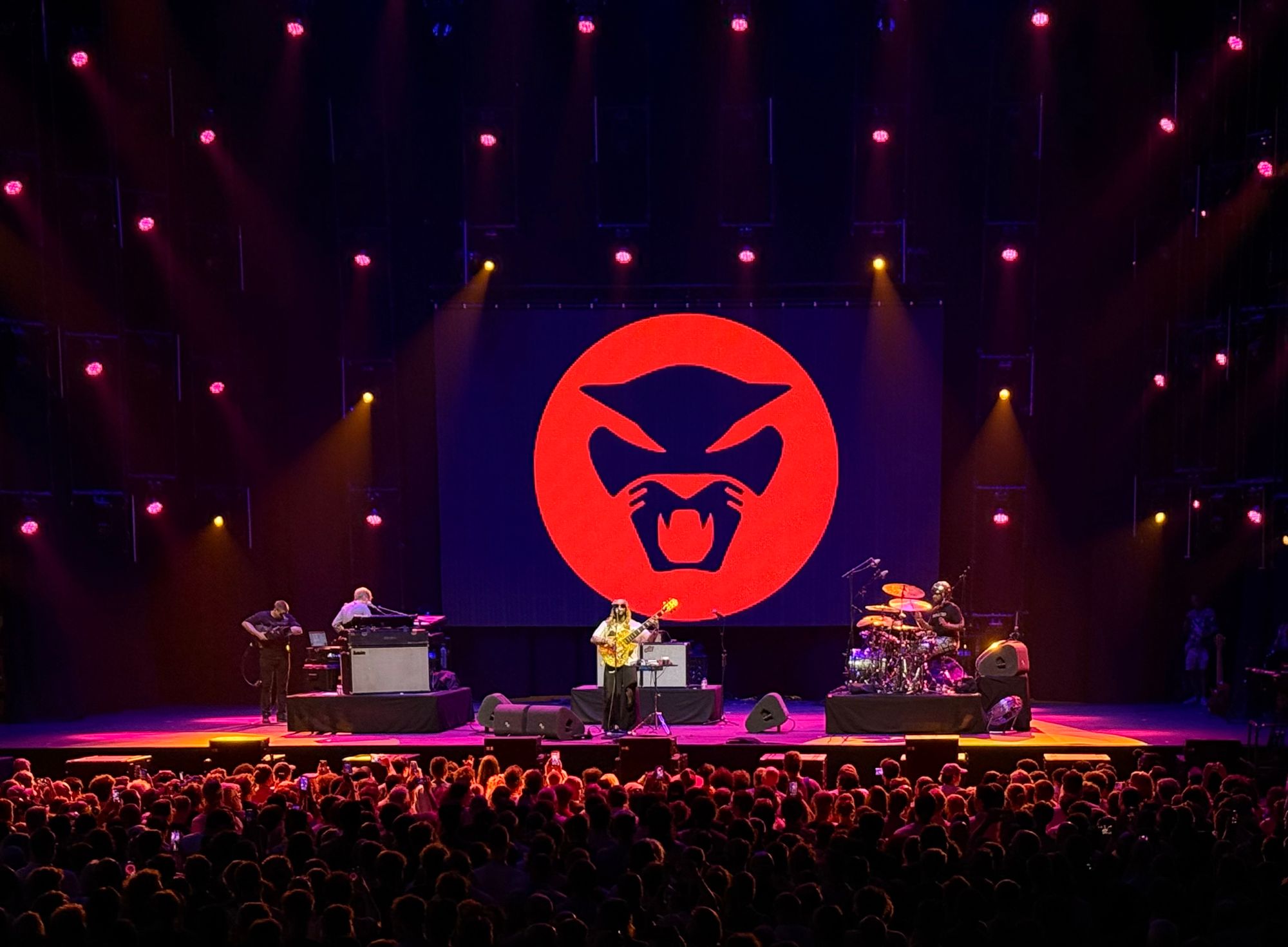 Thundercat playing a golden bass in middle of stage, on his left is someone on synths, on his right a drummer, his logo is in the back