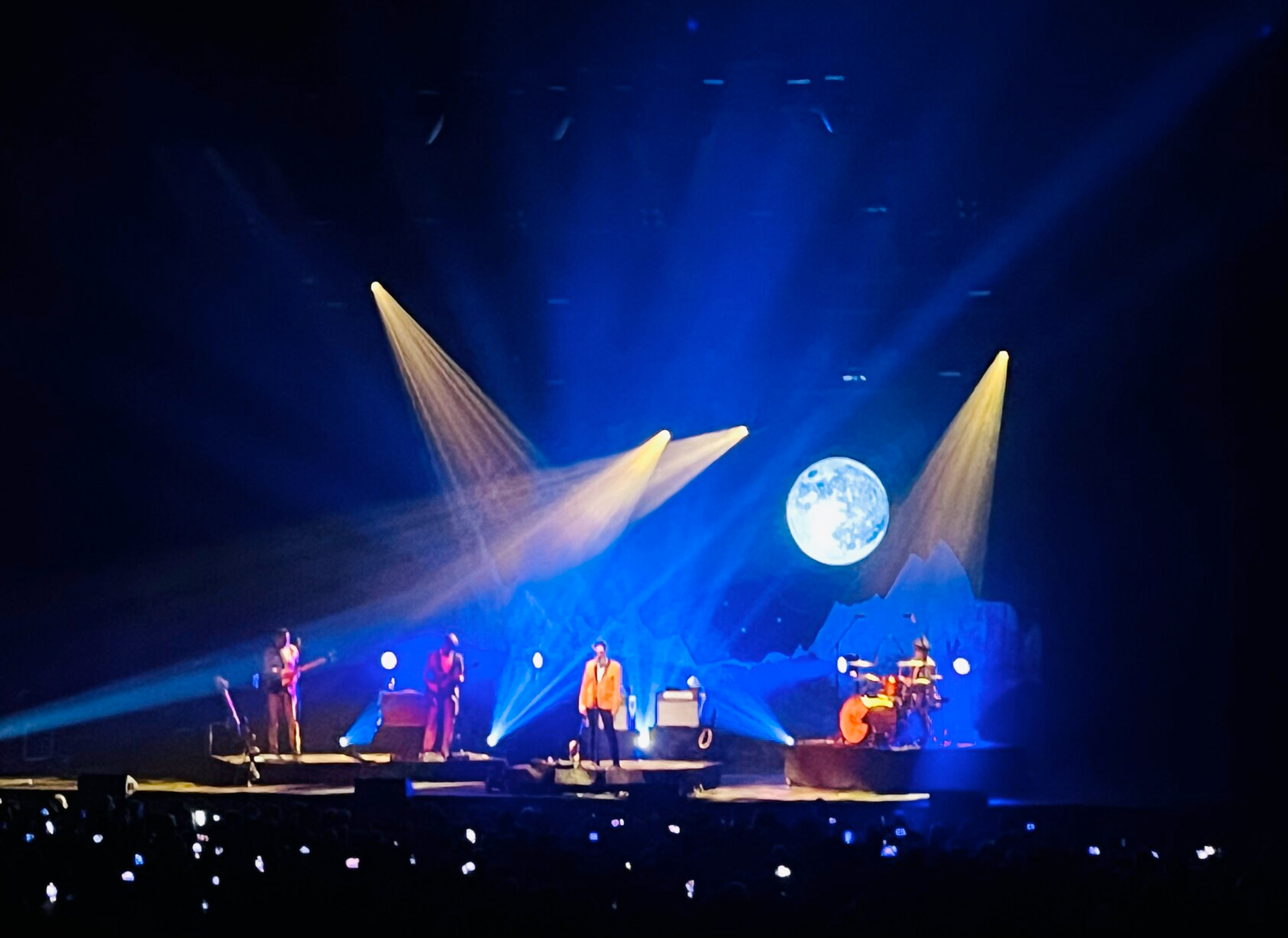 four man band, with front man in orange suit, background lightning is mostly blue with some yellow, and there is a full moon projected on the right