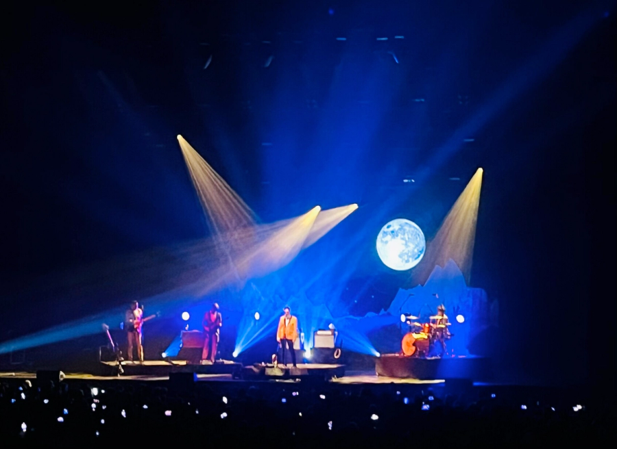 four man band, with front man in orange suit, background lightning is mostly blue with some yellow, and there is a full moon projected on the right