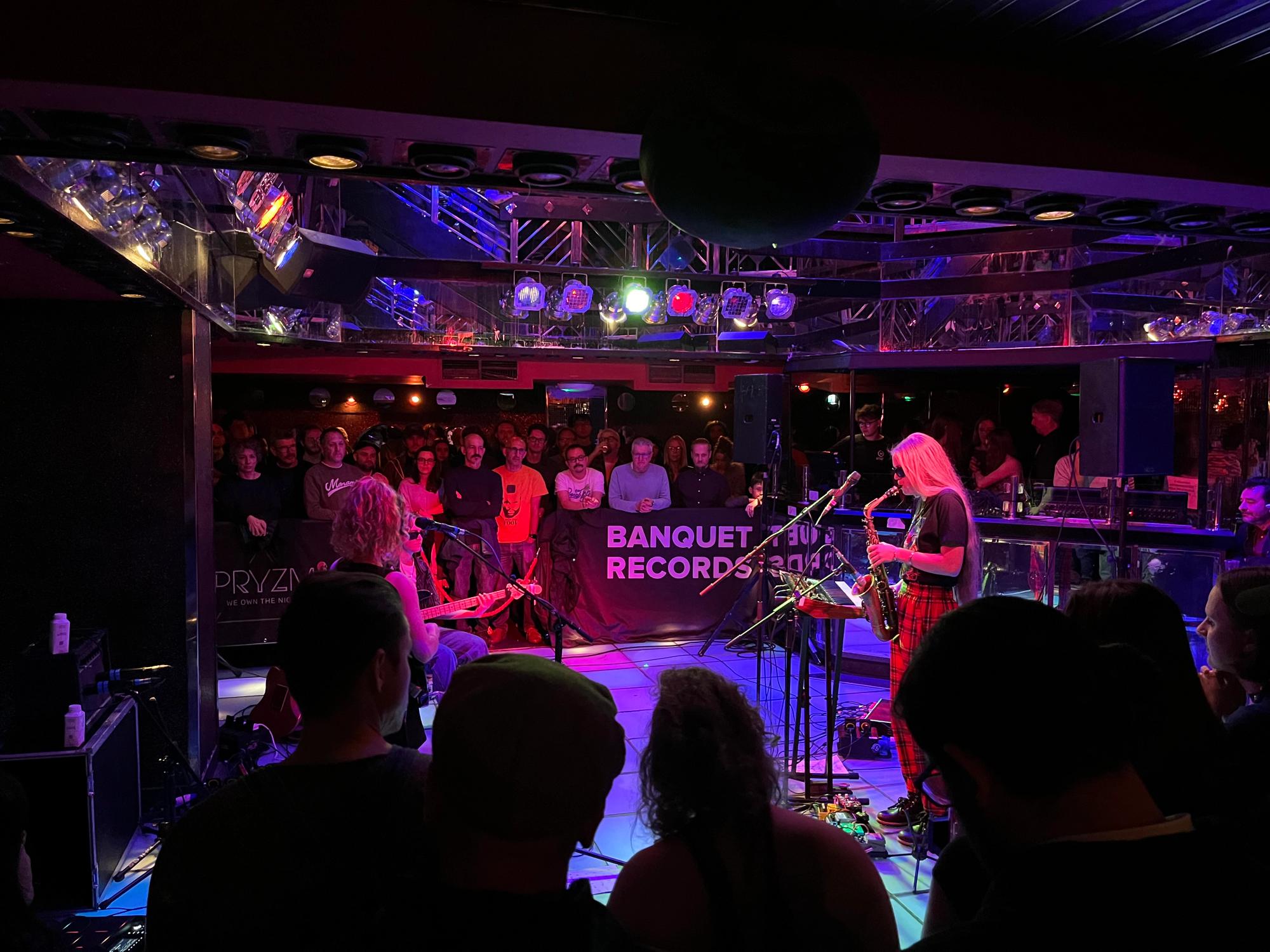 3 artists, one on saxophone, on the left is Nilufer, in a venue that has audience members around the stage