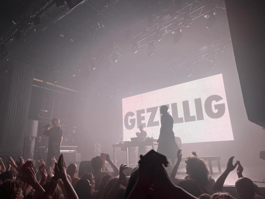 two rappers with their mc on a stage, behind the world gezellig is displayed in huge black letters on a white background. 