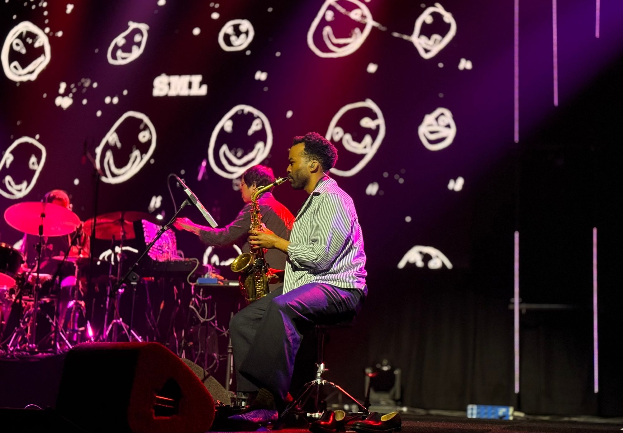 saxophonist looking happy while playing sax, on the back is a lot of children style handdrawn faces and the letters SML