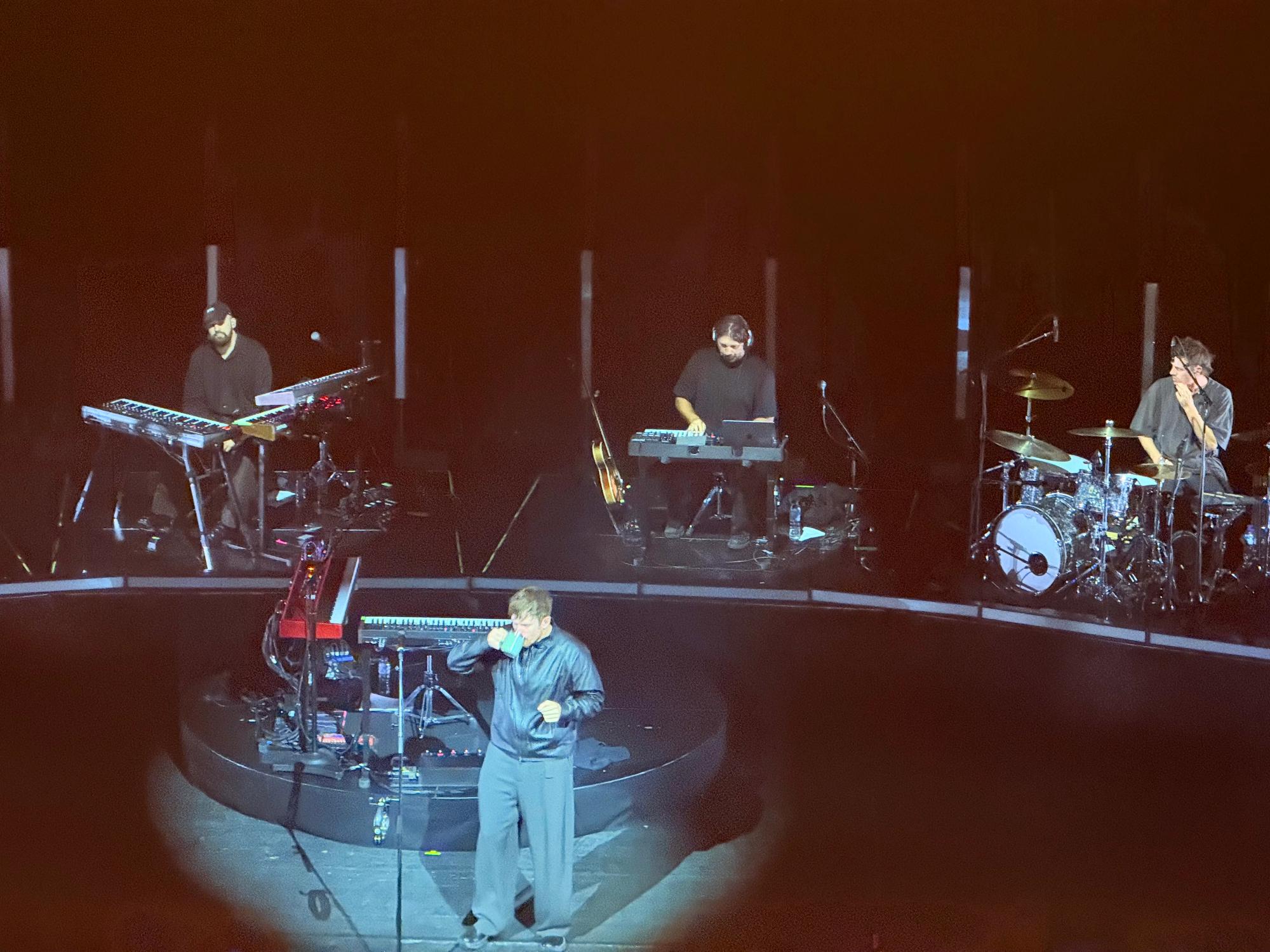 James drinking from a bug, in front of his keyboards, synthesisers and four musicians behind including a drummer and various synths