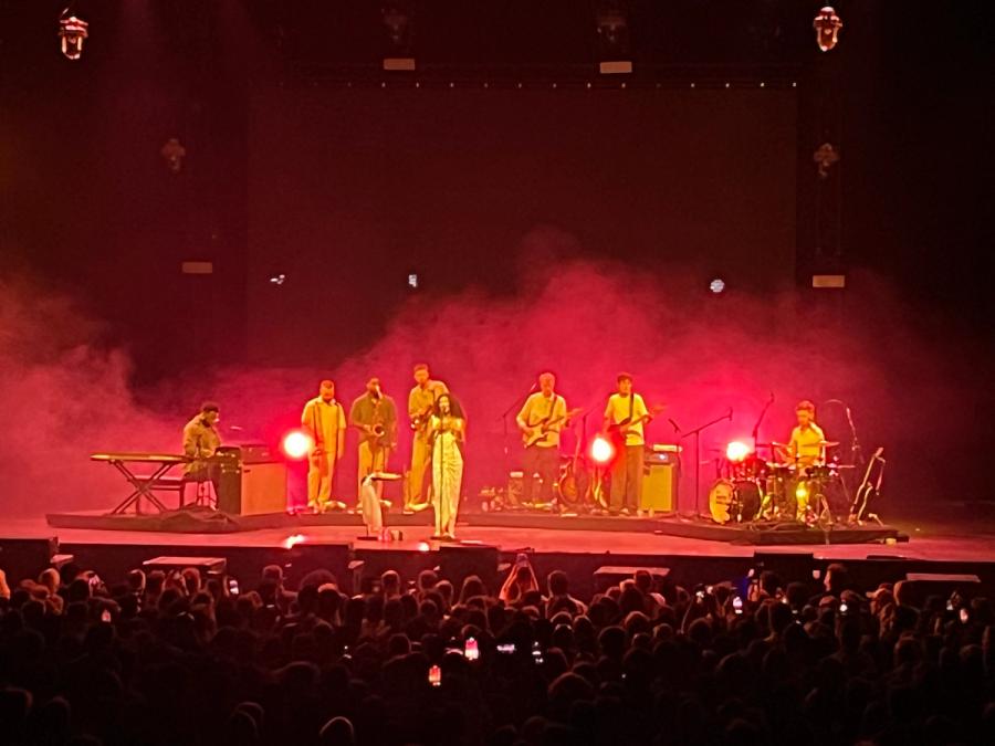 olivia in middle of stage with red lights and audience visible, behind her are pianist, trumpets, bass and drums