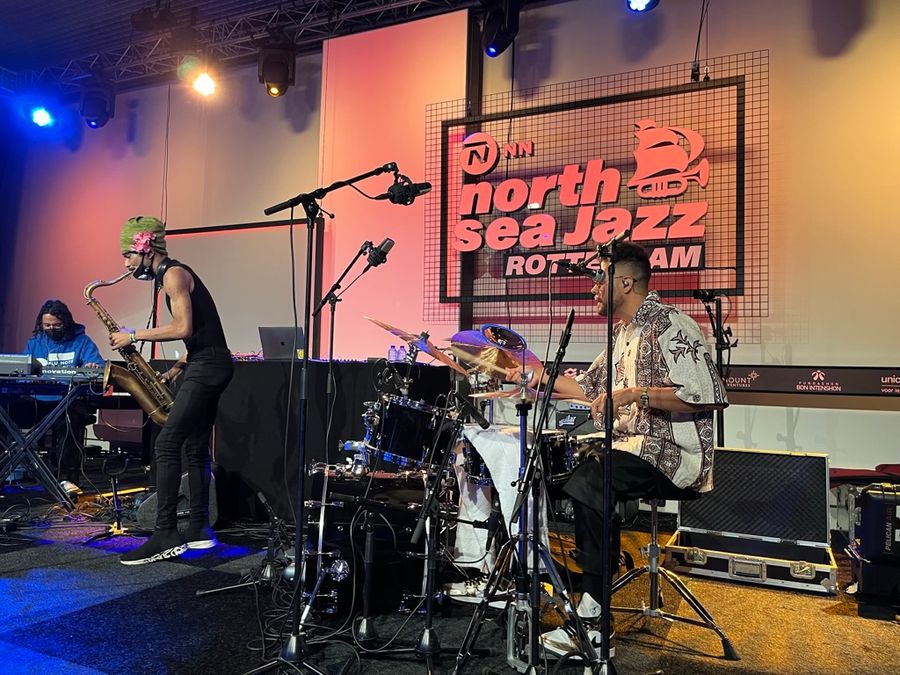 from left to right a keyboard player, saxophonist and drummer, behind them is a large North Sea Jazz Rotterdam sign with the iconic trumpet/ship logo.