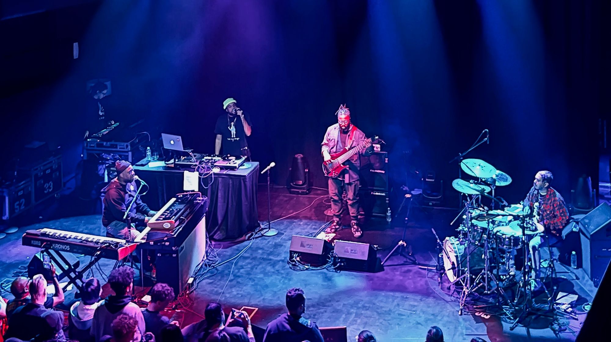 stage with from left to right Robert Glasper playing a digital piano and talking into a microphone, his MC, a bass player and a drummer