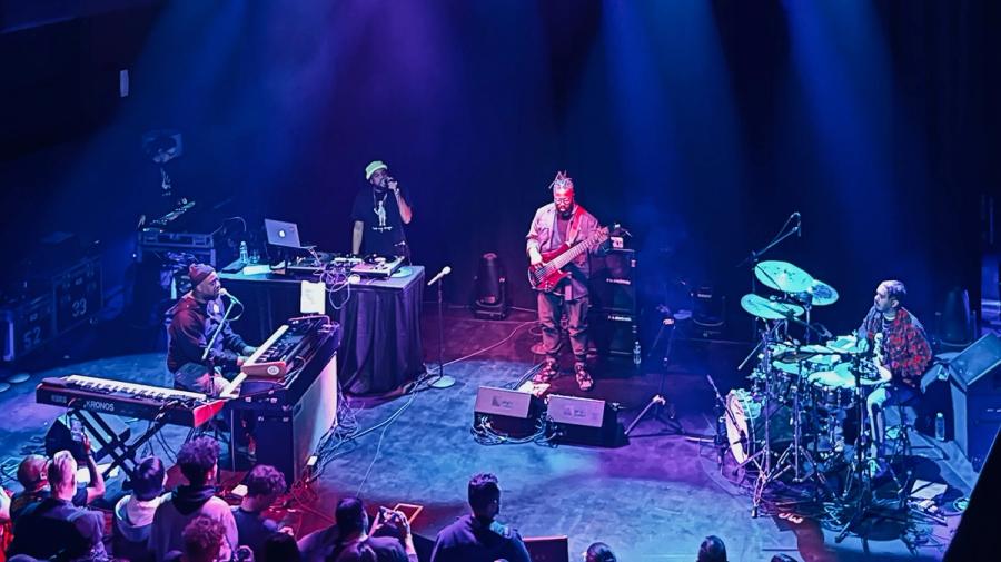 stage with from left to right Robert Glasper playing a digital piano and talking into a microphone, his MC, a bass player and a drummer