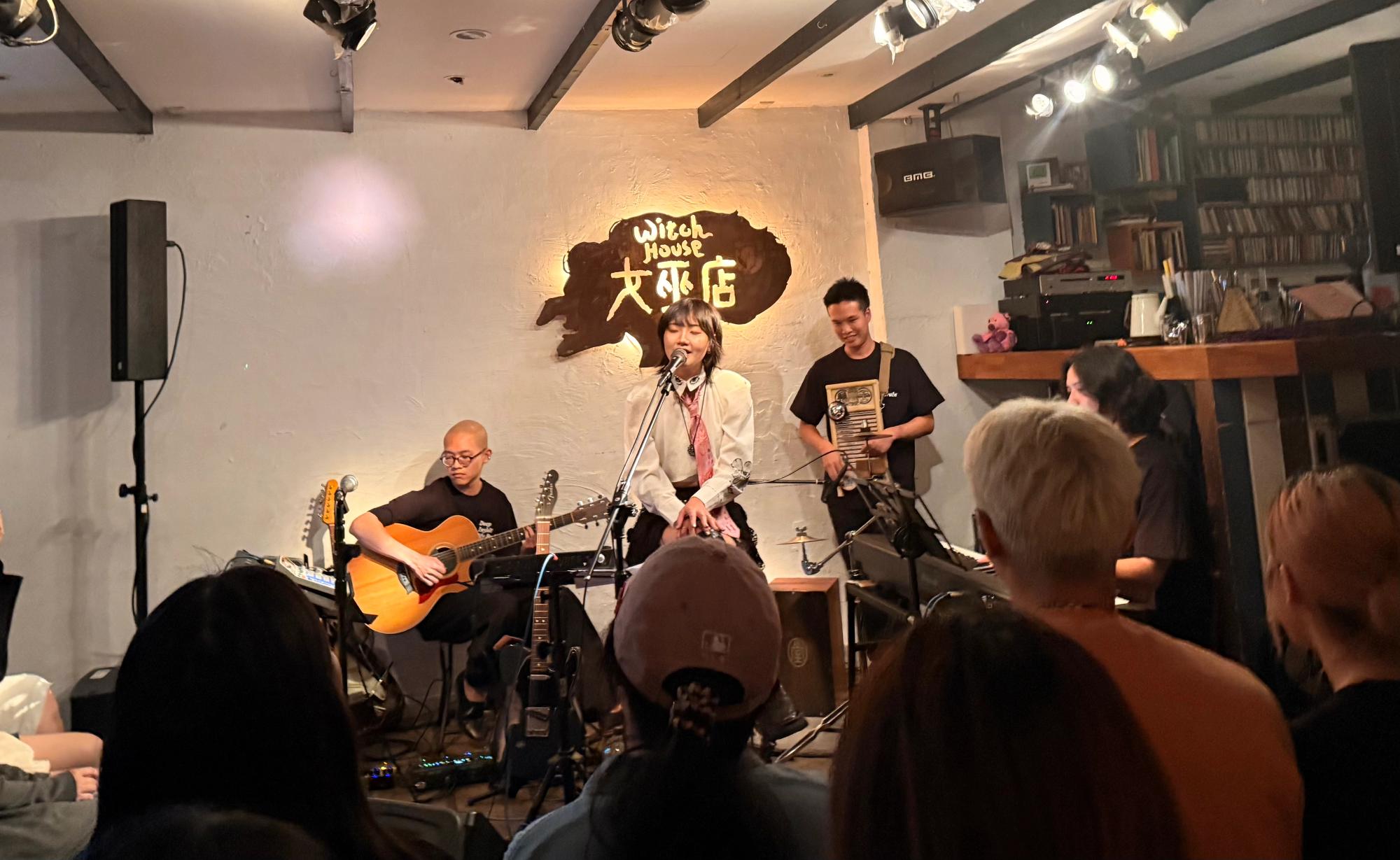 artist on chair, left of her is her guitarist, drums on right and lots of CDs visible in bookcase behind bad
