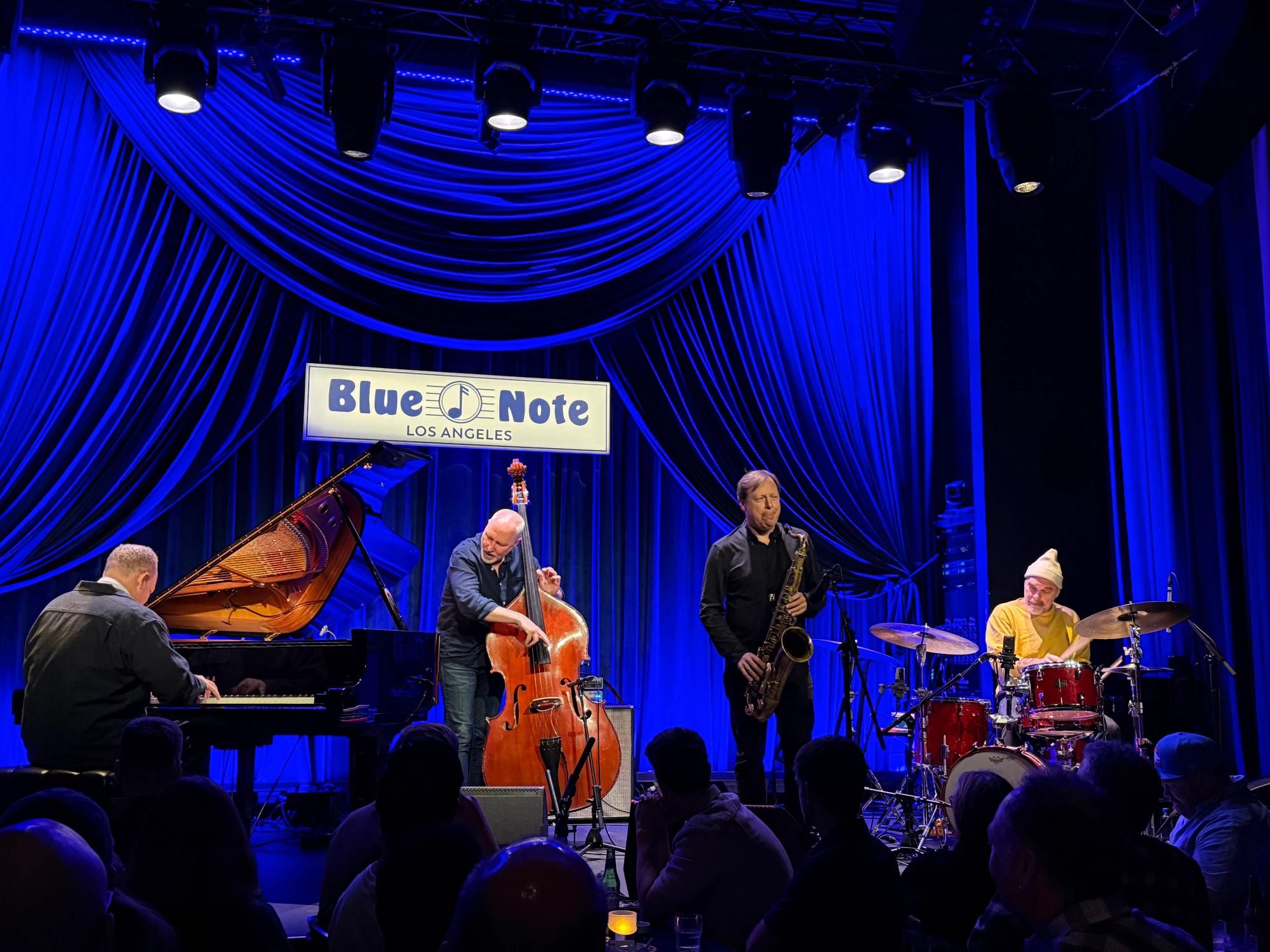 Craig Taborn, Reid Anderson, Chris Potter and Dave King on stage playing grand piano, double bass, saxophone and drums, in the back is a blue curtain and a blue note los angeles sign