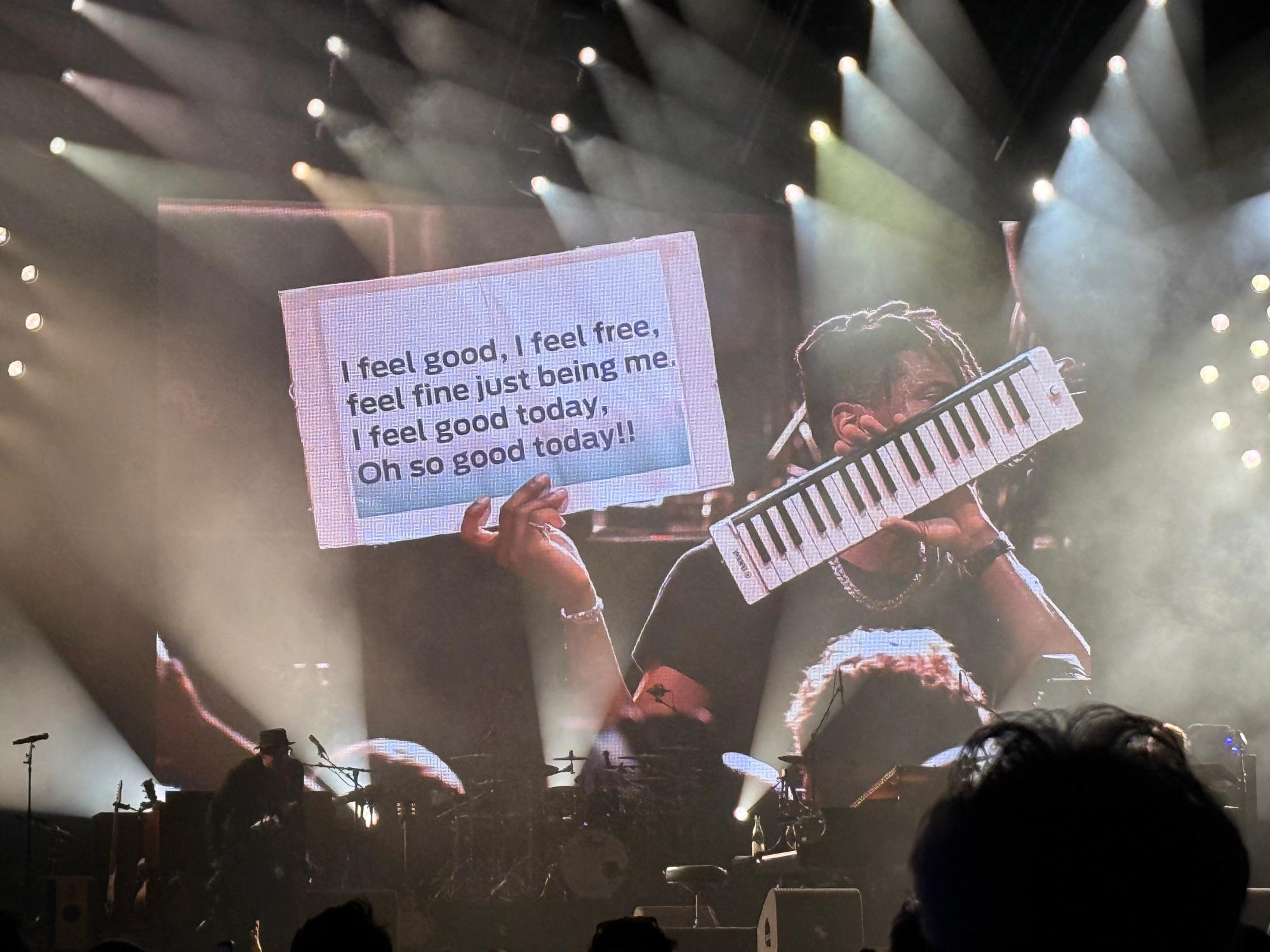 Jon Batiste holding a tiny keyboard in one hand and in the other a sign that says I feel good, I feel free, feel fine just being me, I feel good today, oh so good today