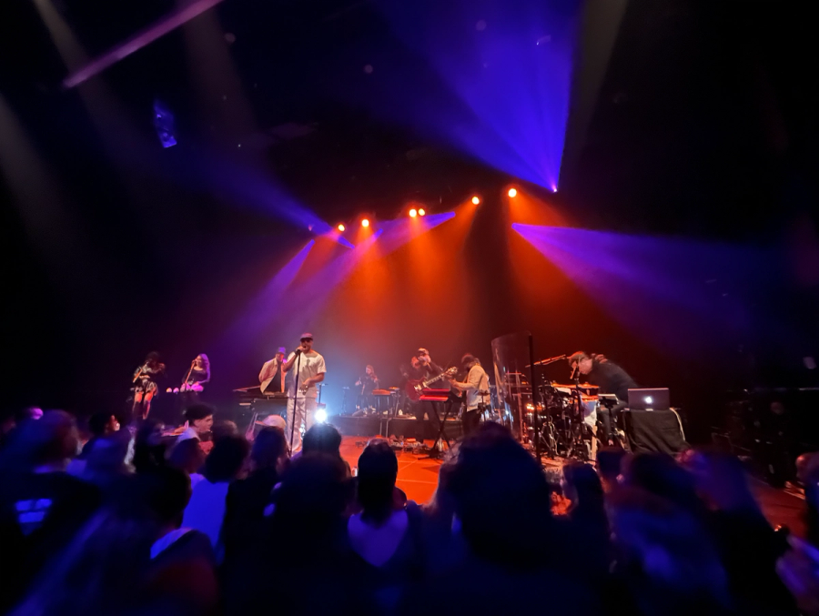 rapper wearing hat on stage, with two female backing vocals on the left and two violonists,  a guitar player and a drummer on the right