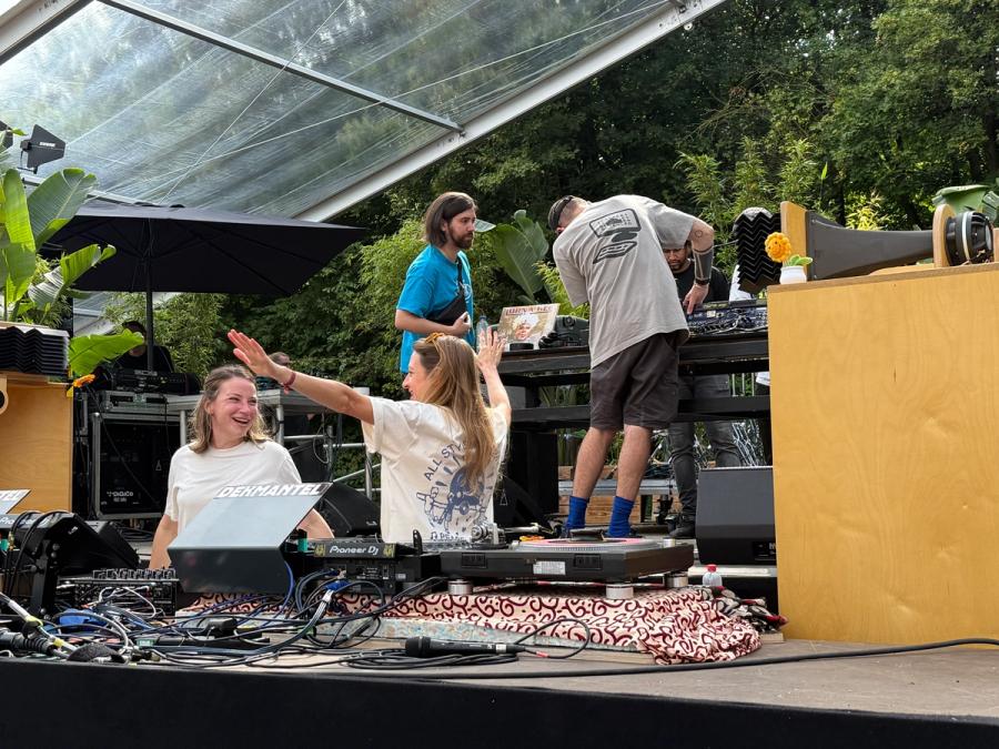 Team Javybz behind turn tables, dancing and laughing, behind them people are setting up for next set