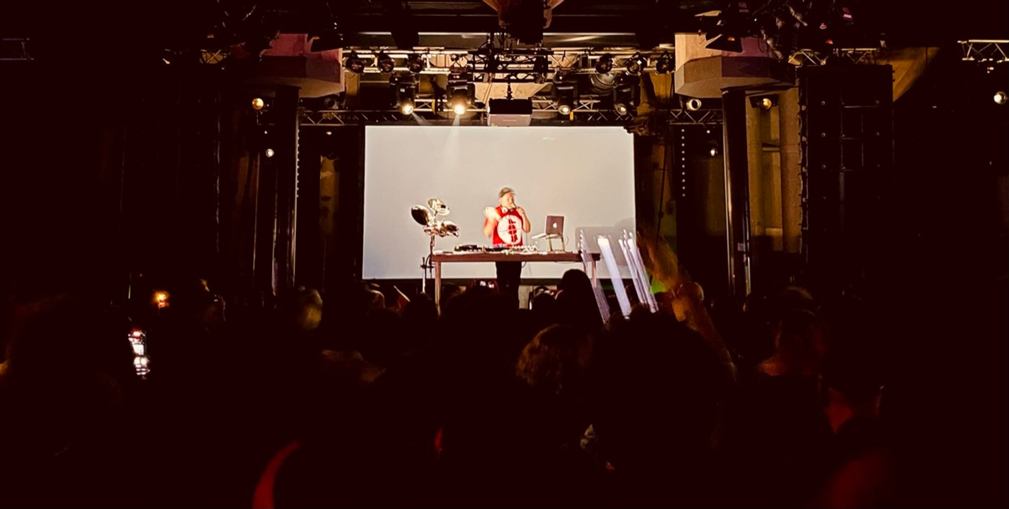 Man holding mic behind a table with turntables and a laptop, as seen from crowd