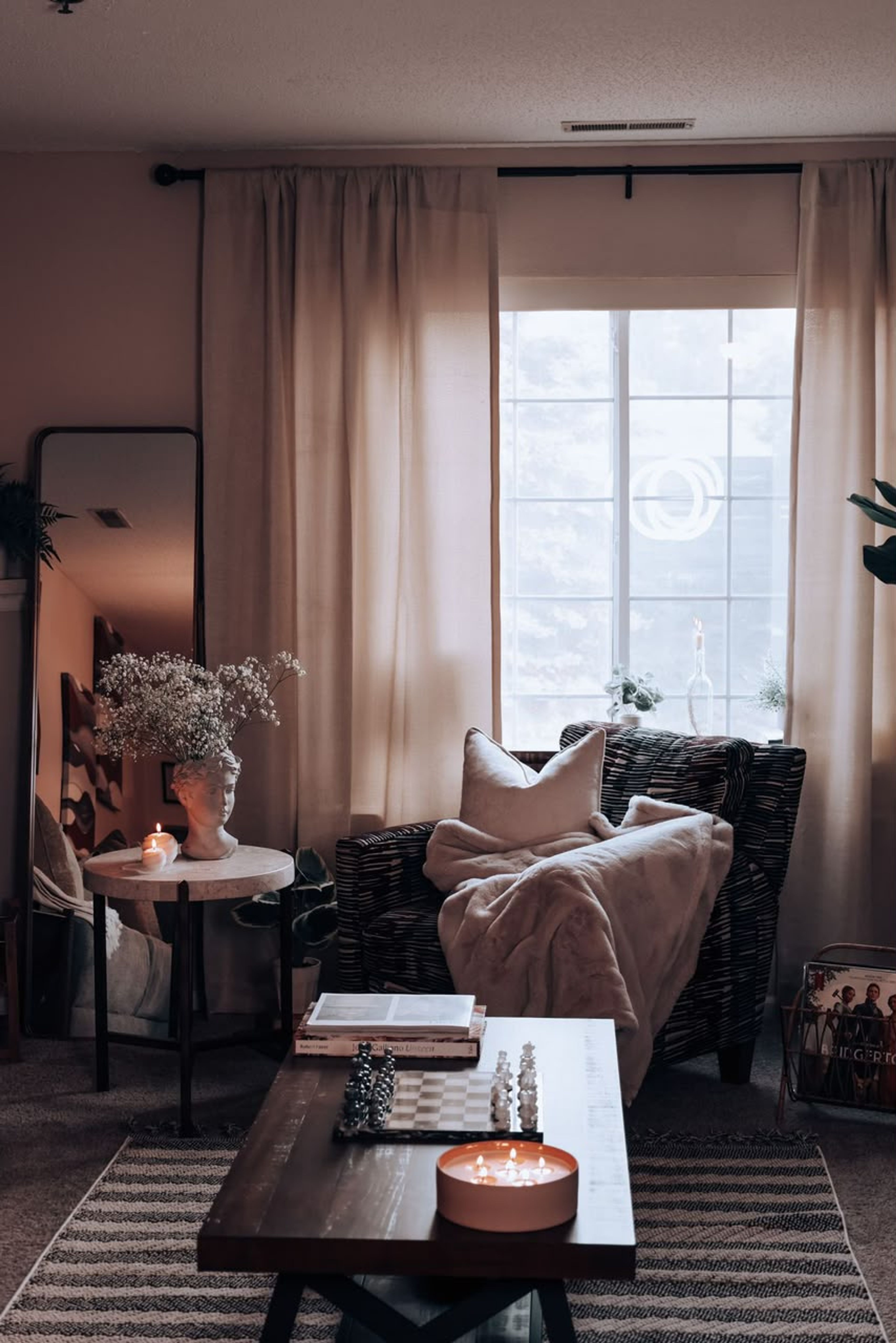 A softly lit living room with linen curtains, an armchair with a cream throw, a dark wood coffee table with a chess set and candle, and a sculptural bust vase of dried flowers on a side table